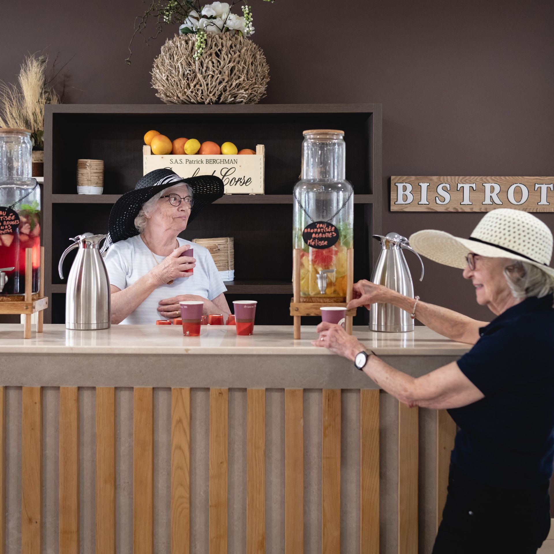 Deux femmes âgées portant des chapeaux dans un bar-bistro, l'une verse un verre tandis que l'autre tient une tasse.