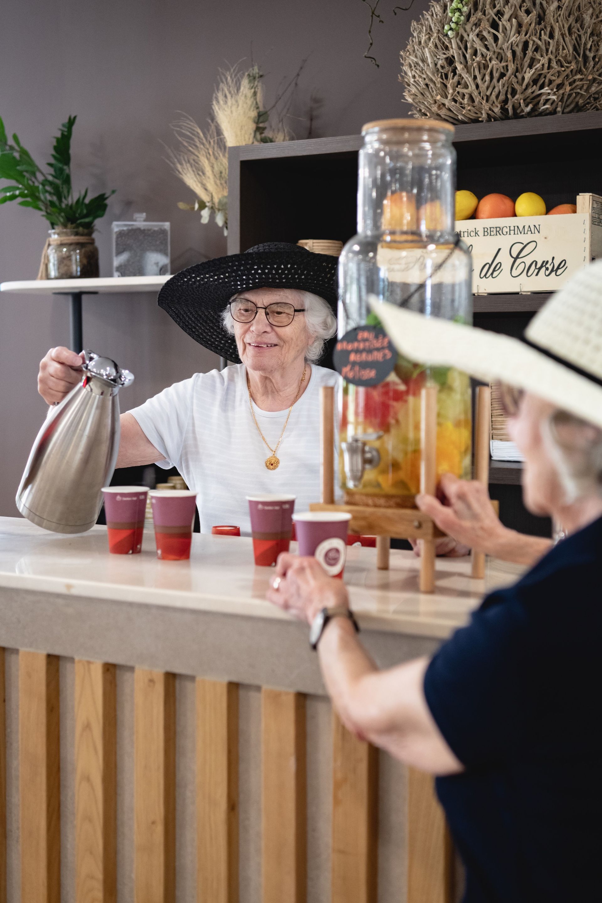 Une femme portant un chapeau verse une boisson à une autre femme à un comptoir avec un distributeur rempli de fruits.