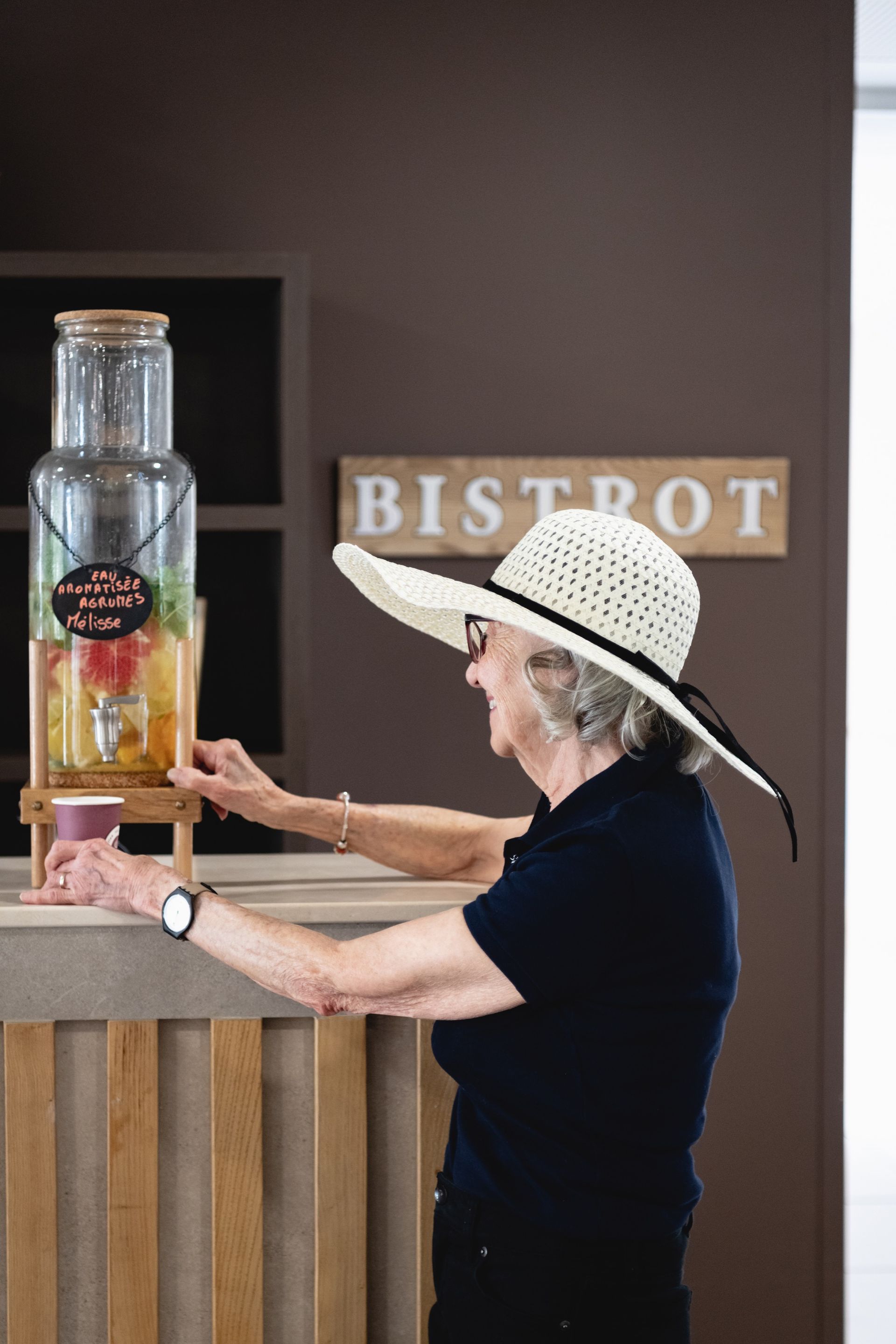 Femme portant un chapeau de soleil dans un bistrot, remplissant une tasse à partir d'un distributeur d'eau infusée aux fruits.