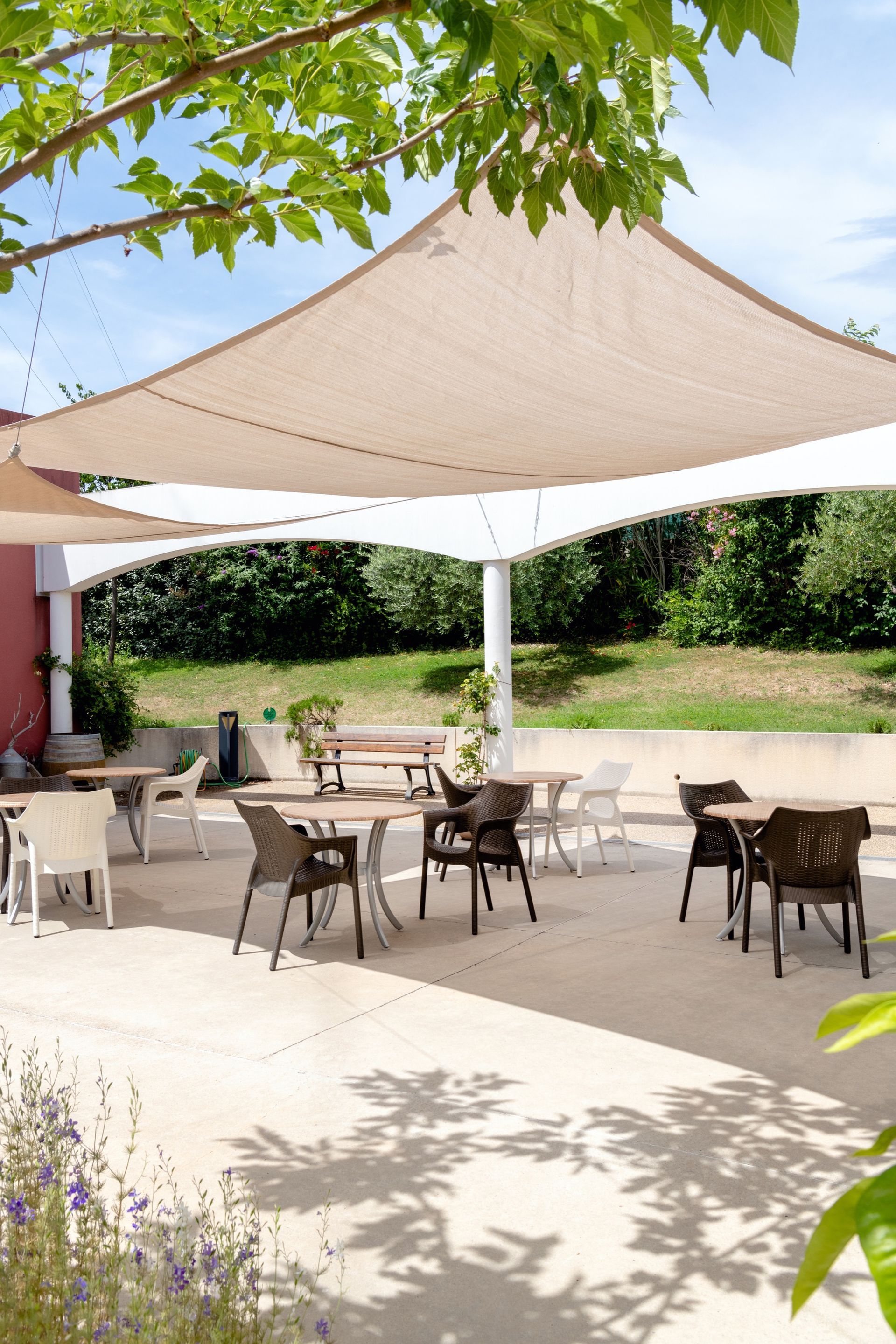 Espace repas extérieur avec tables et chaises sous un abat-jour beige, entouré de verdure.