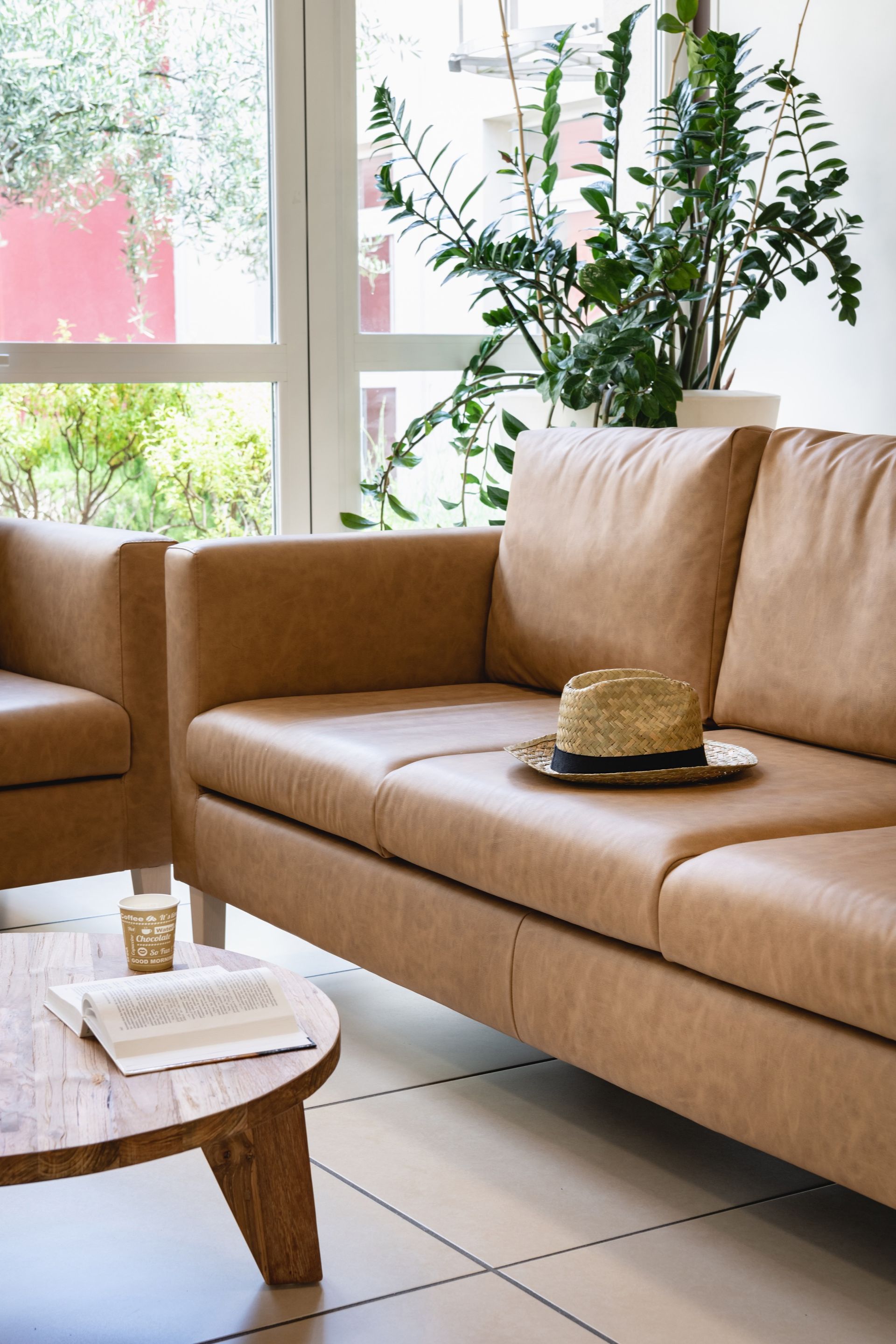 Canapé et fauteuil en cuir beige, chapeau de paille et table ronde en bois près d'une fenêtre.
