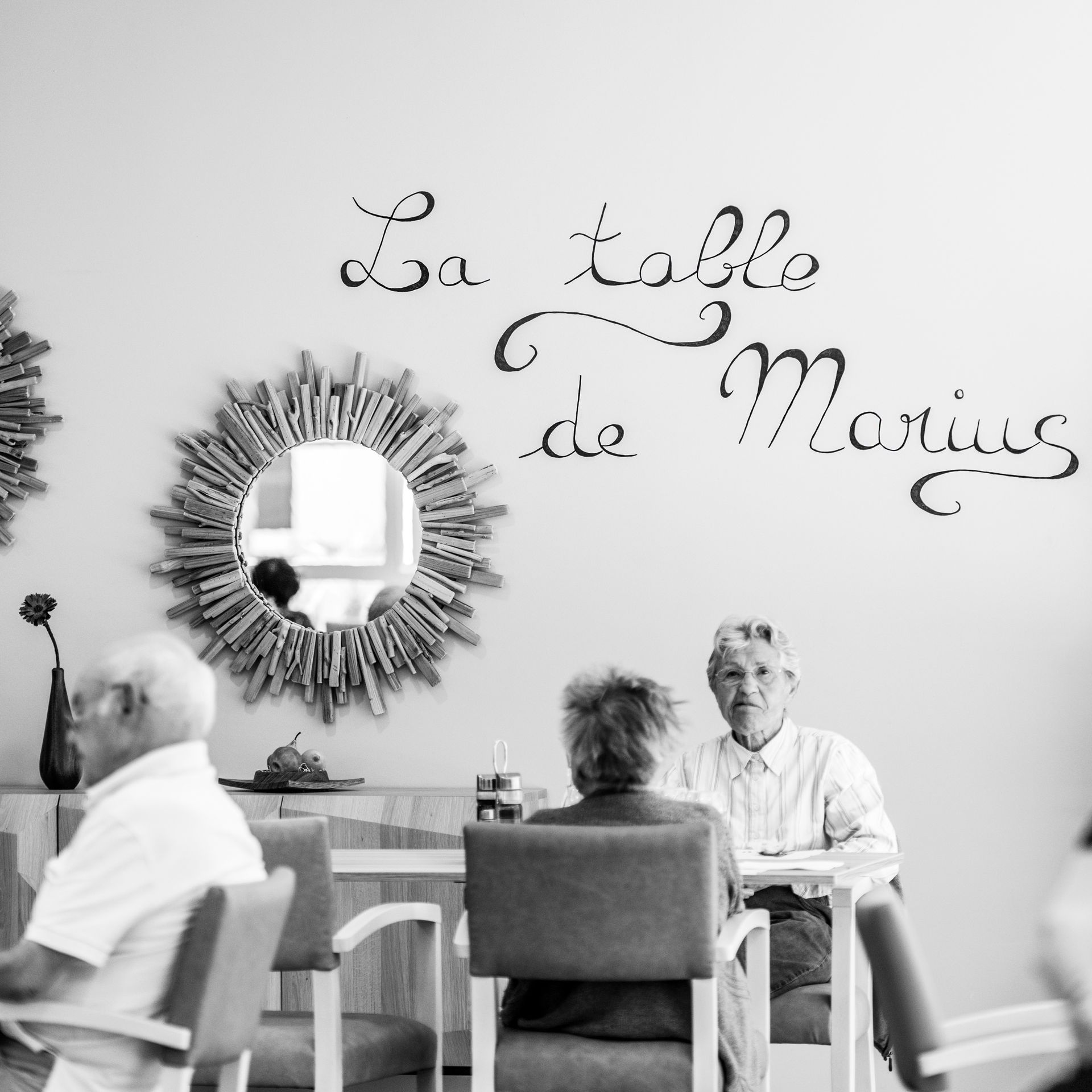 Des personnes âgées assises à une table à manger dans une pièce ; le texte sur le mur indique « La table de Marius ».