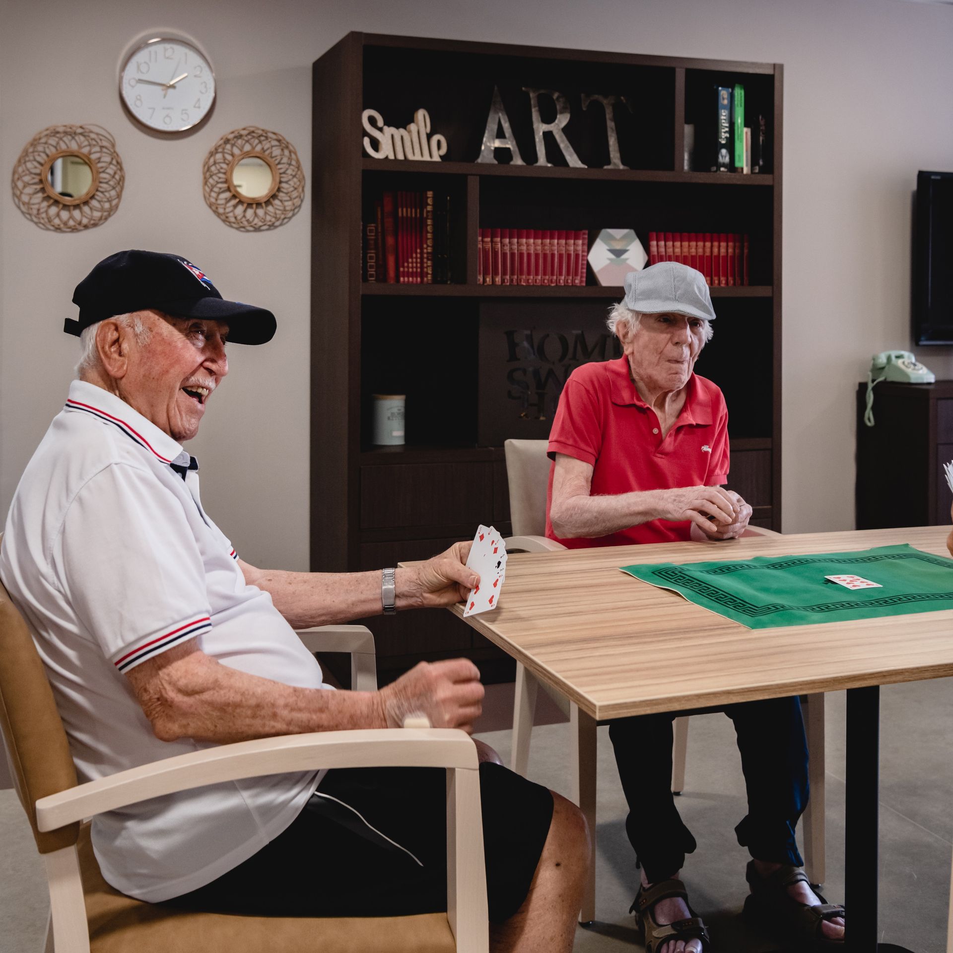Deux hommes âgés jouent aux cartes à une table dans une pièce. L'un d'eux sourit, coiffé d'une casquette de baseball.