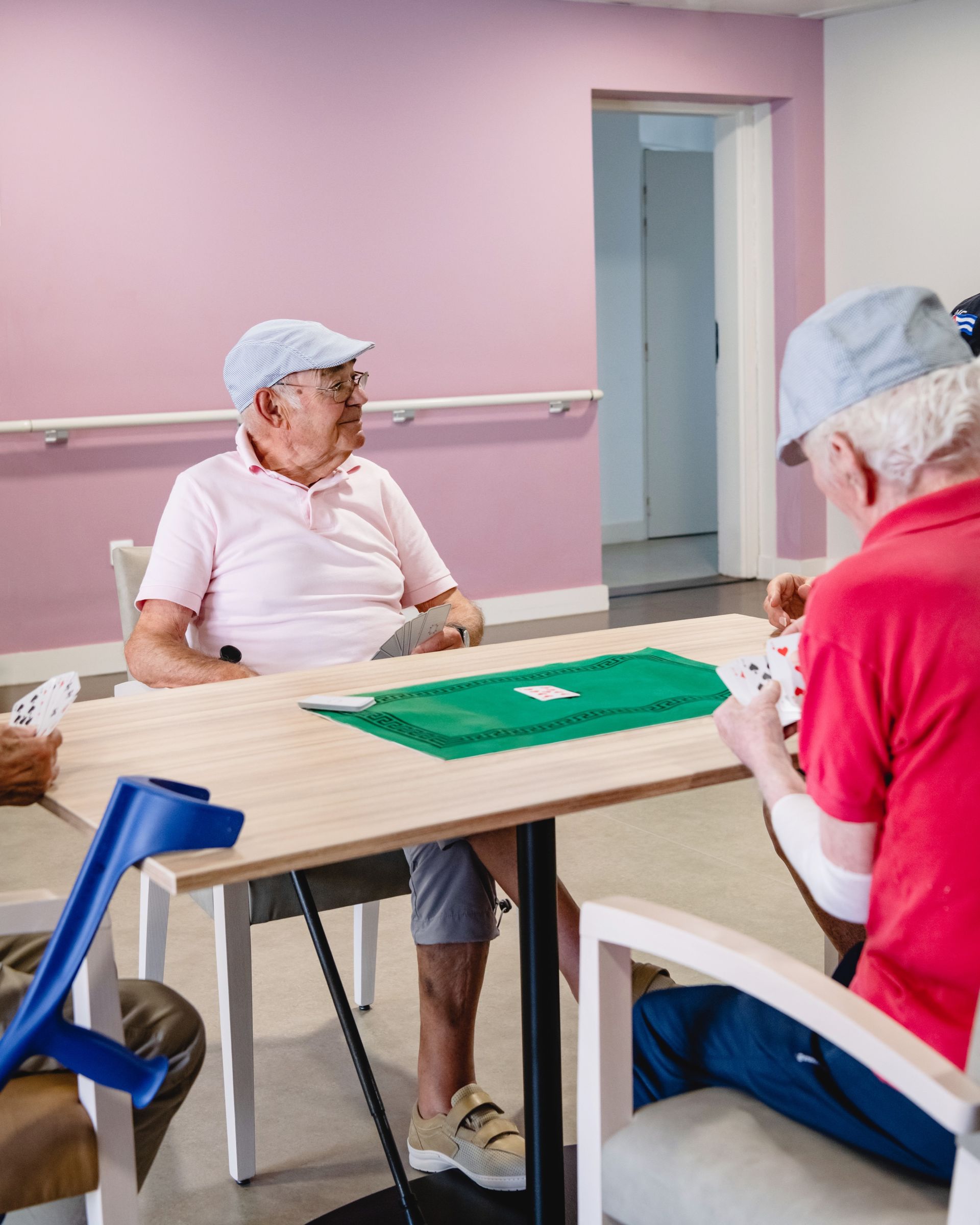 Deux hommes âgés jouent aux cartes à une table dans une pièce. L'un porte un chapeau et une chemise rose, l'autre une chemise rouge.