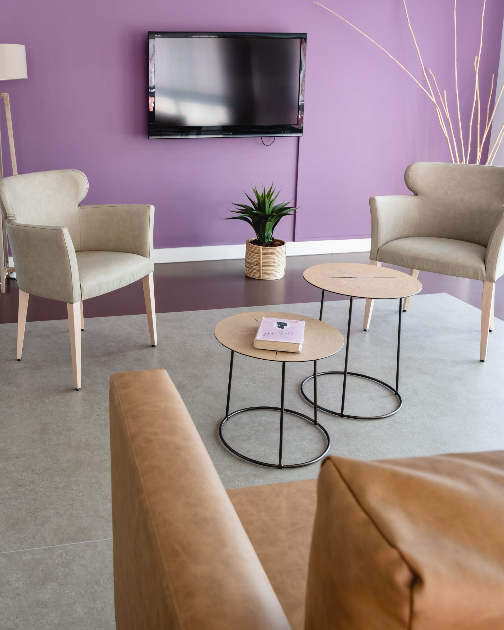 Salon avec mur violet, deux fauteuils, deux petites tables, une télé et une plante en pot.