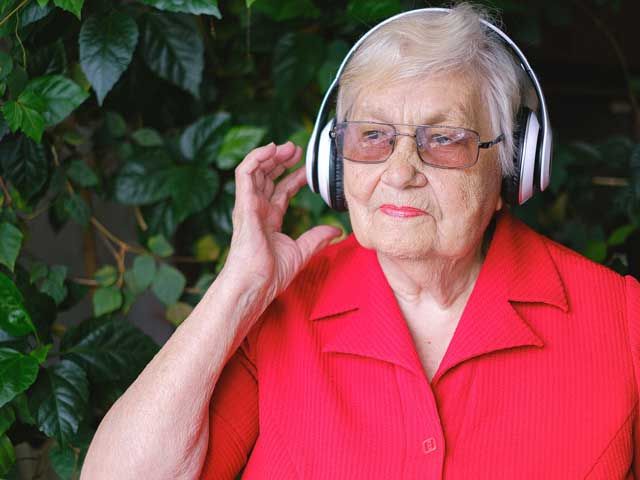 Personne âgée portant un casque audio, ajustant l'écouteur ; chemise rouge, lunettes, feuillage vert.