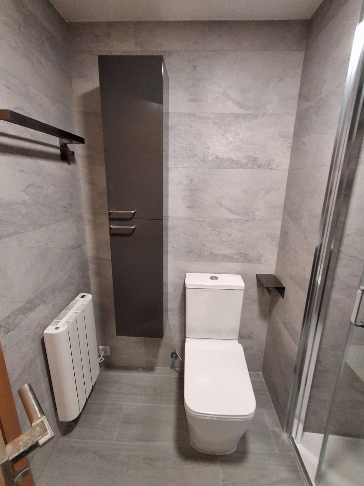 Baño pequeño y moderno con inodoro, ducha de cristal, radiador toallero y paredes alicatadas en gris.