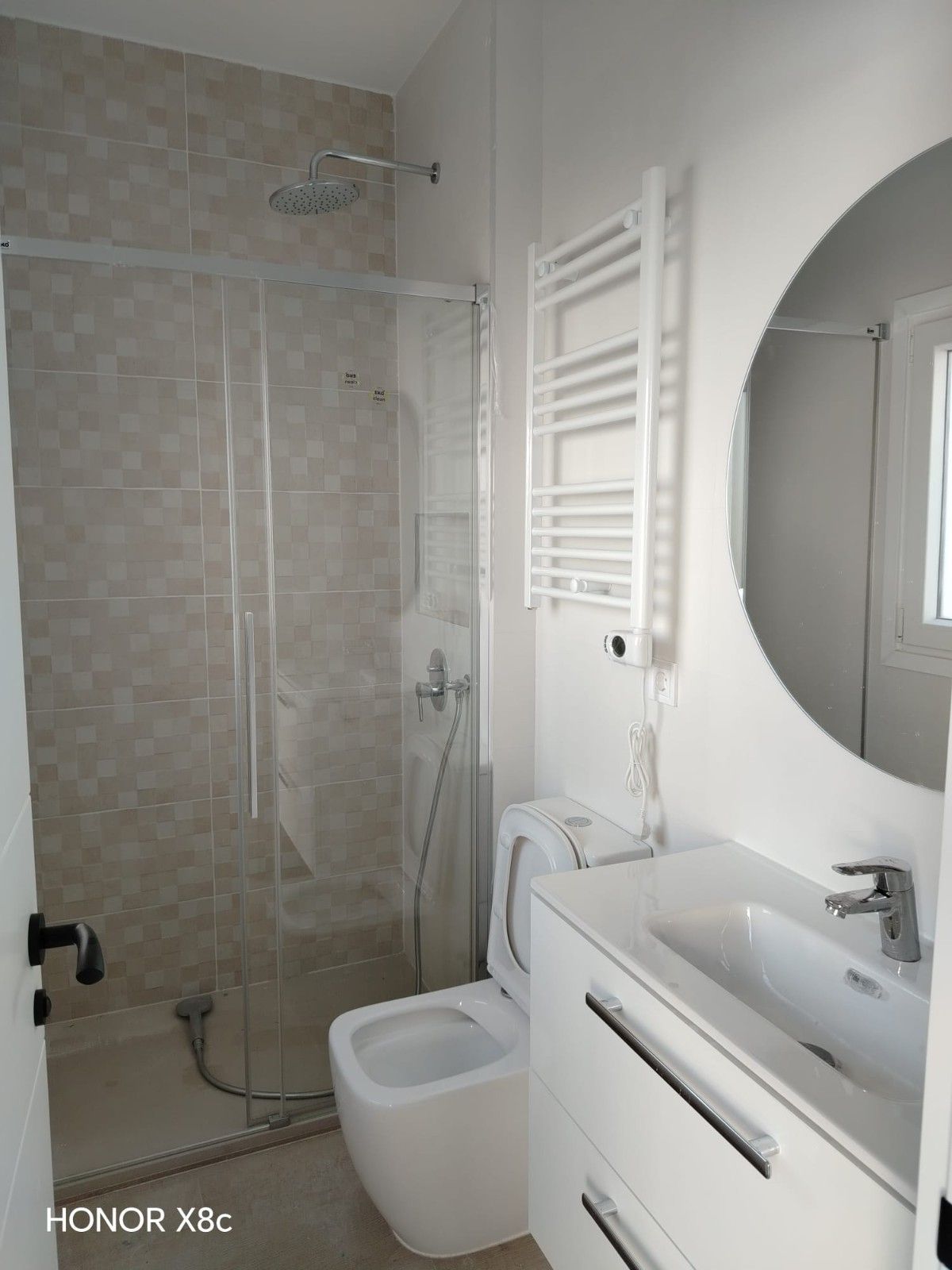 Baño moderno con ducha de cristal, inodoro suspendido, lavabo blanco, espejo redondo y radiador toallero.