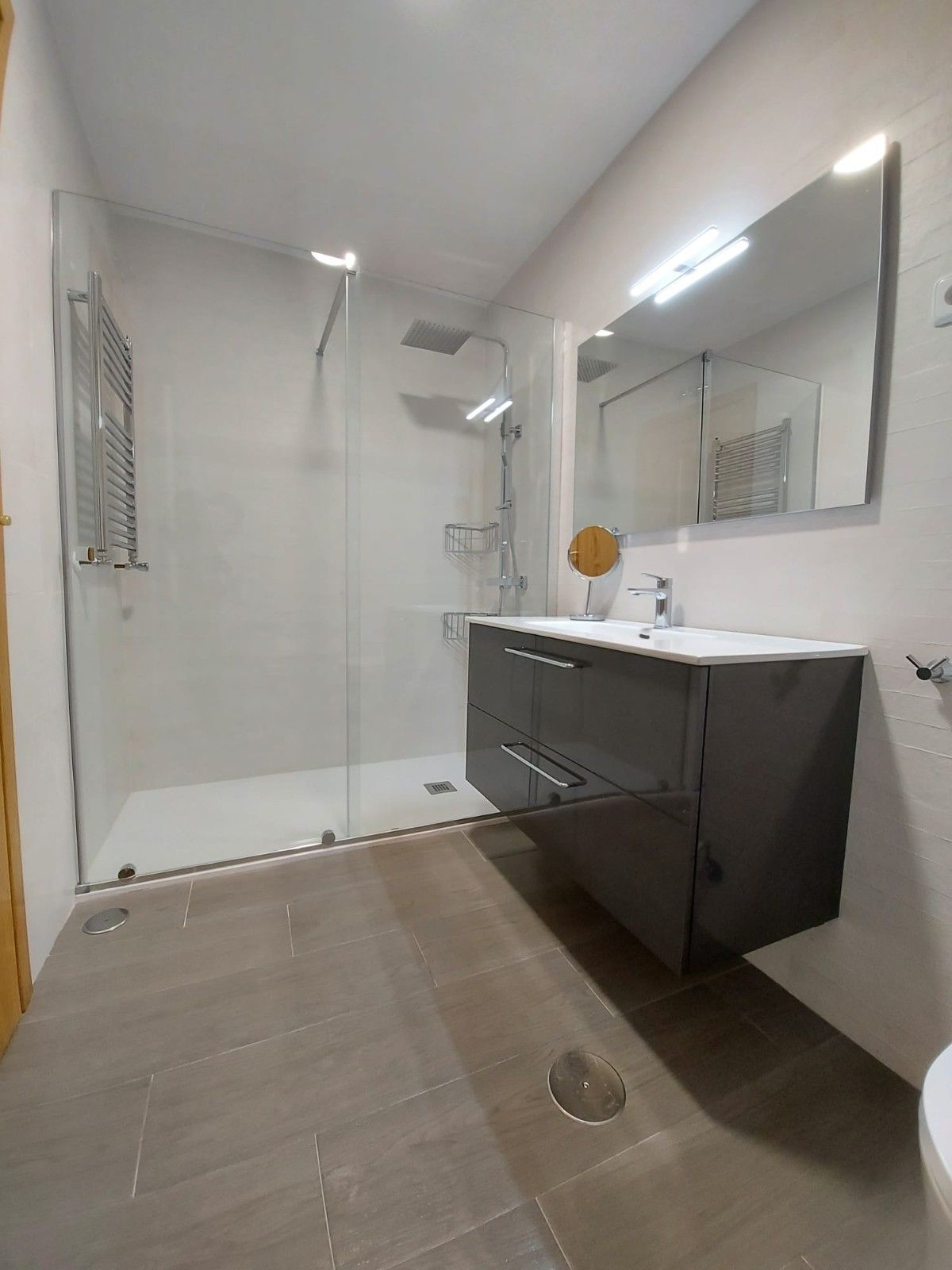 Baño moderno con ducha de cristal, lavabo suspendido oscuro, espejo y suelo de baldosas.
