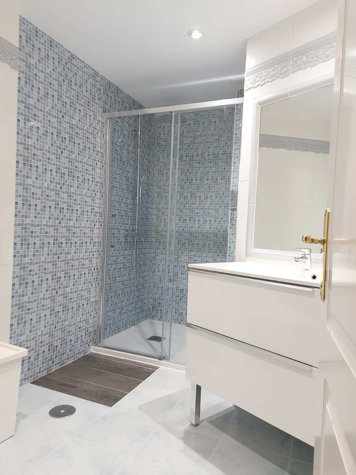 Baño moderno con ducha revestida de azulejos azules, lavabo blanco, espejo y alfombrilla gris.