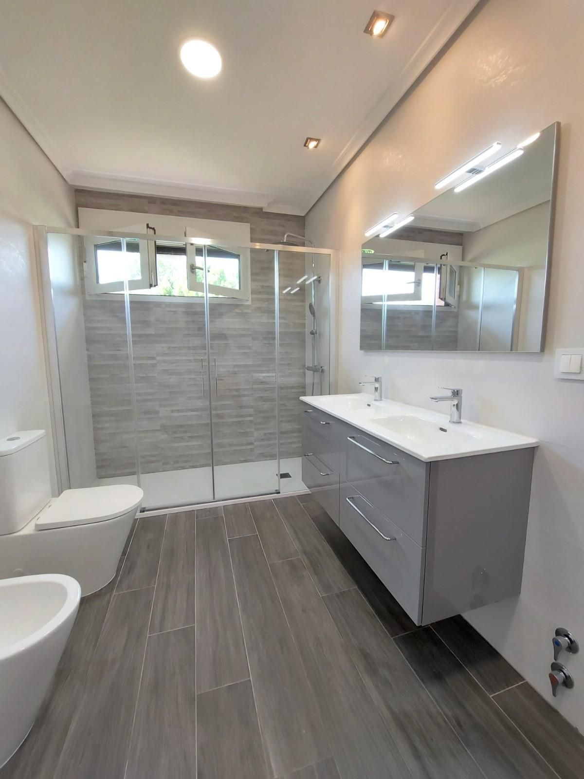 Baño moderno con suelo de baldosas grises, ducha de cristal, tocador con doble lavabo y espejo grande.