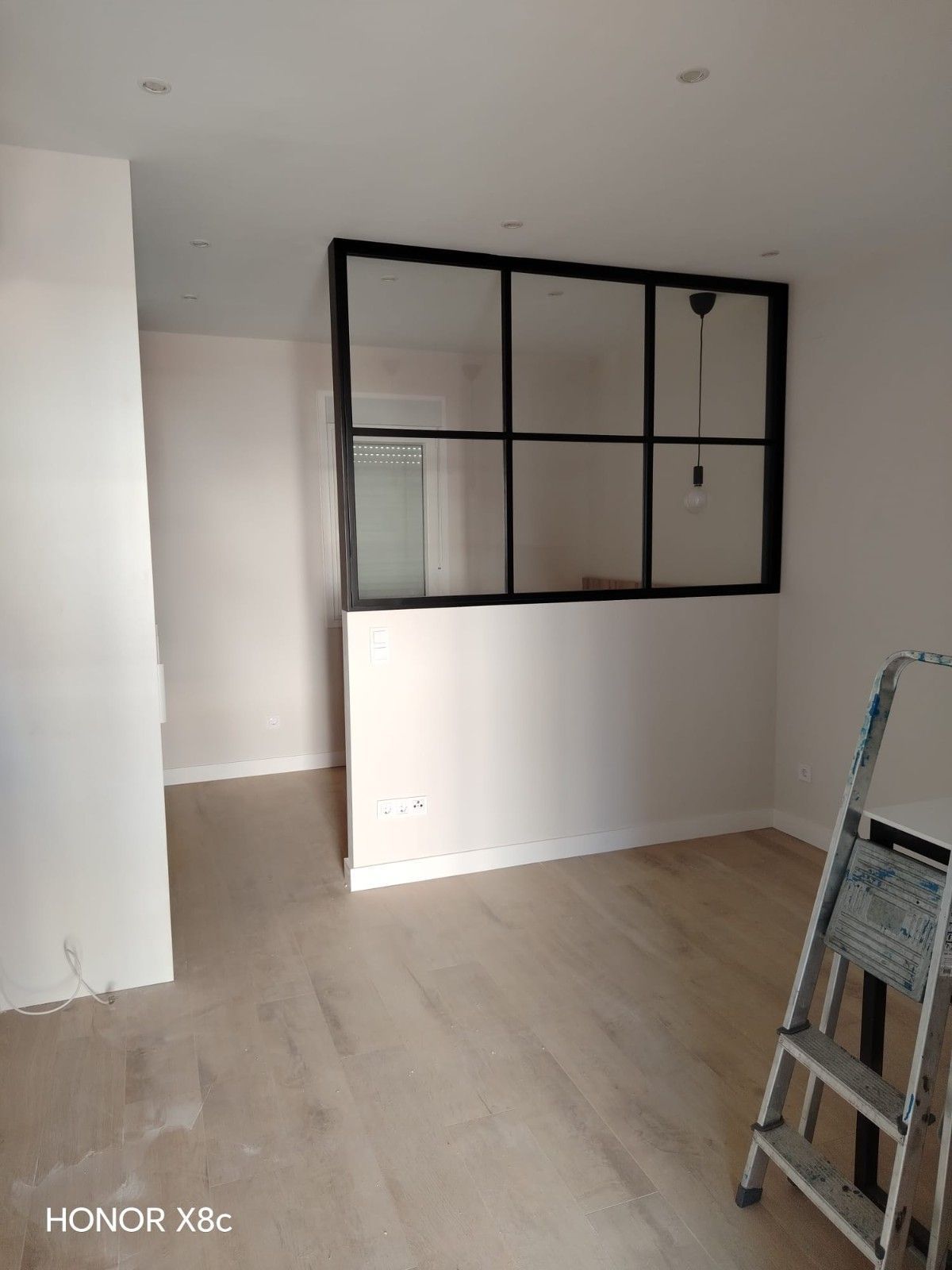 Habitación vacía con suelo de madera clara, paredes blancas, ventana divisoria con marco negro y una escalera a la derecha.