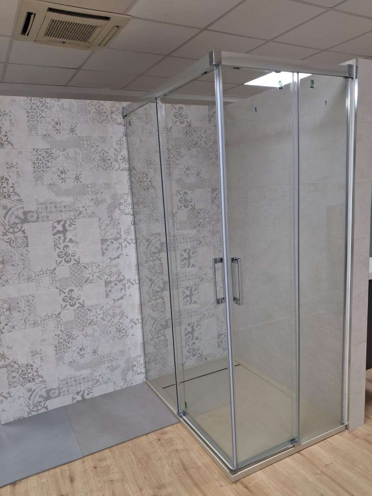 Mampara de ducha de cristal en un baño alicatado con alfombra gris y suelo de imitación madera.