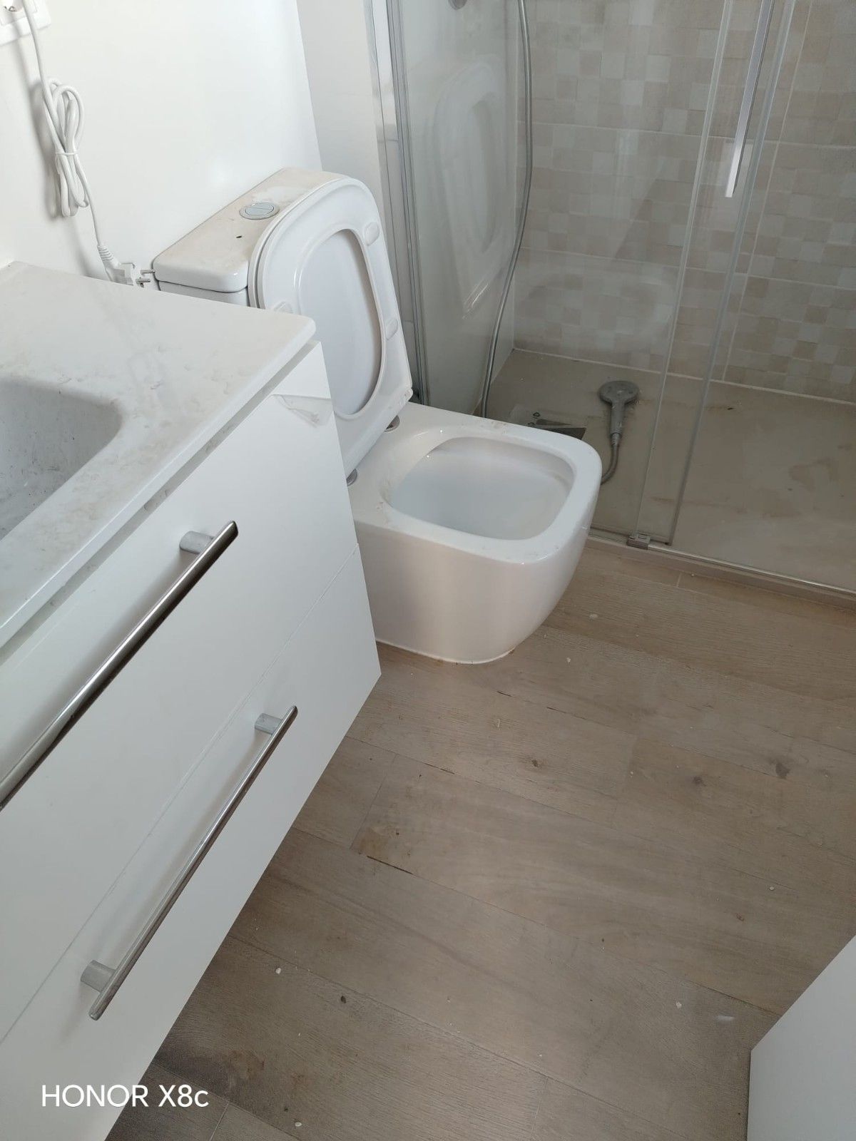 Baño moderno con lavabo blanco, inodoro, bidé y cabina de ducha de cristal sobre suelo de madera clara.