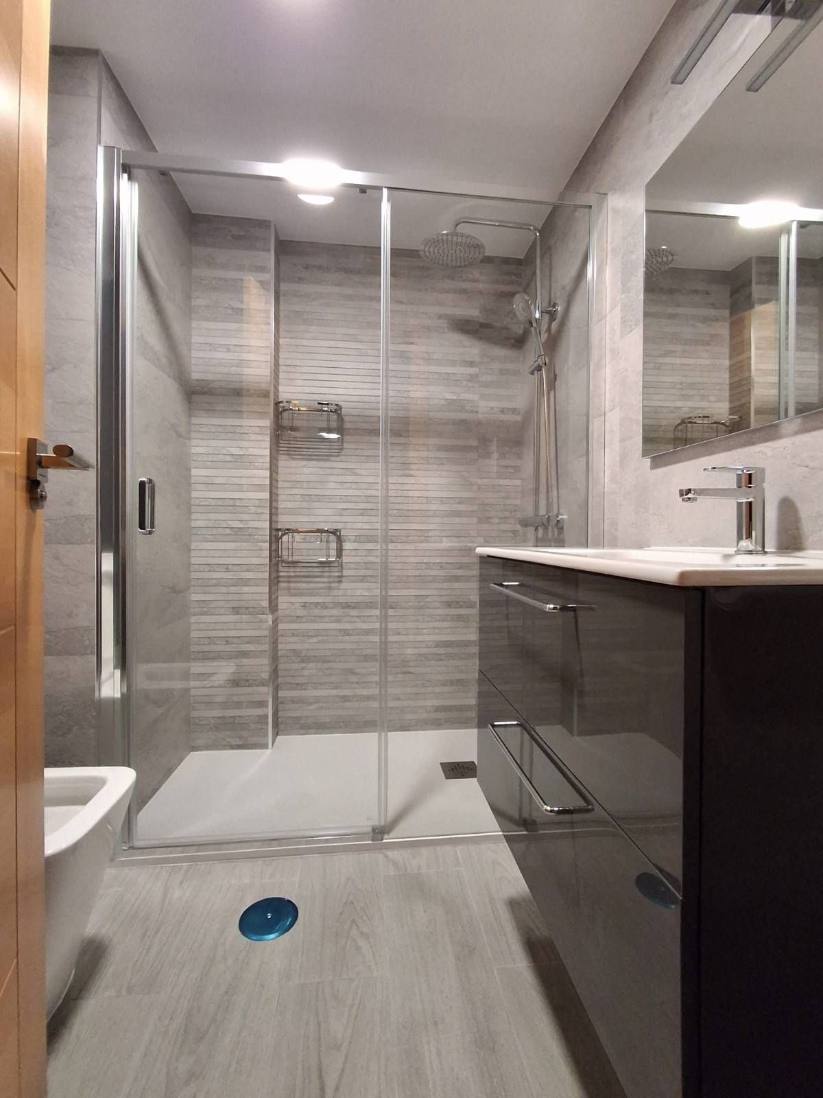 Baño moderno con azulejos grises, ducha de cristal, tocador, espejo e inodoro parcialmente visible.