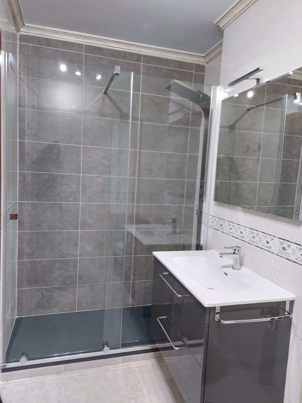 Baño moderno con ducha de azulejos grises, puerta de cristal y lavabo blanco con espejo.