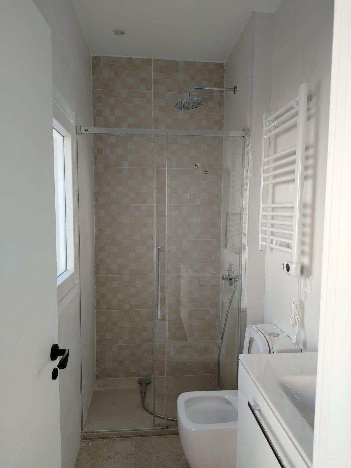 Baño estrecho con ducha, inodoro, lavabo y toallero eléctrico en un pequeño espacio alicatado.
