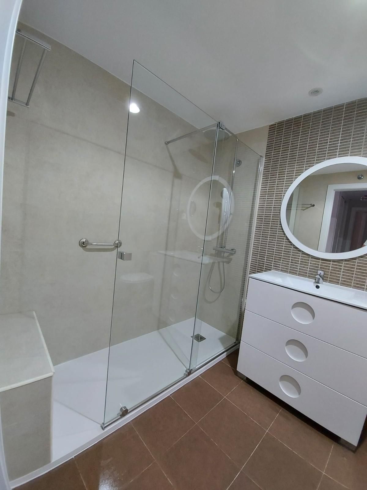 Baño moderno con ducha de cristal, lavabo blanco, espejo redondo y suelo de baldosas marrones.