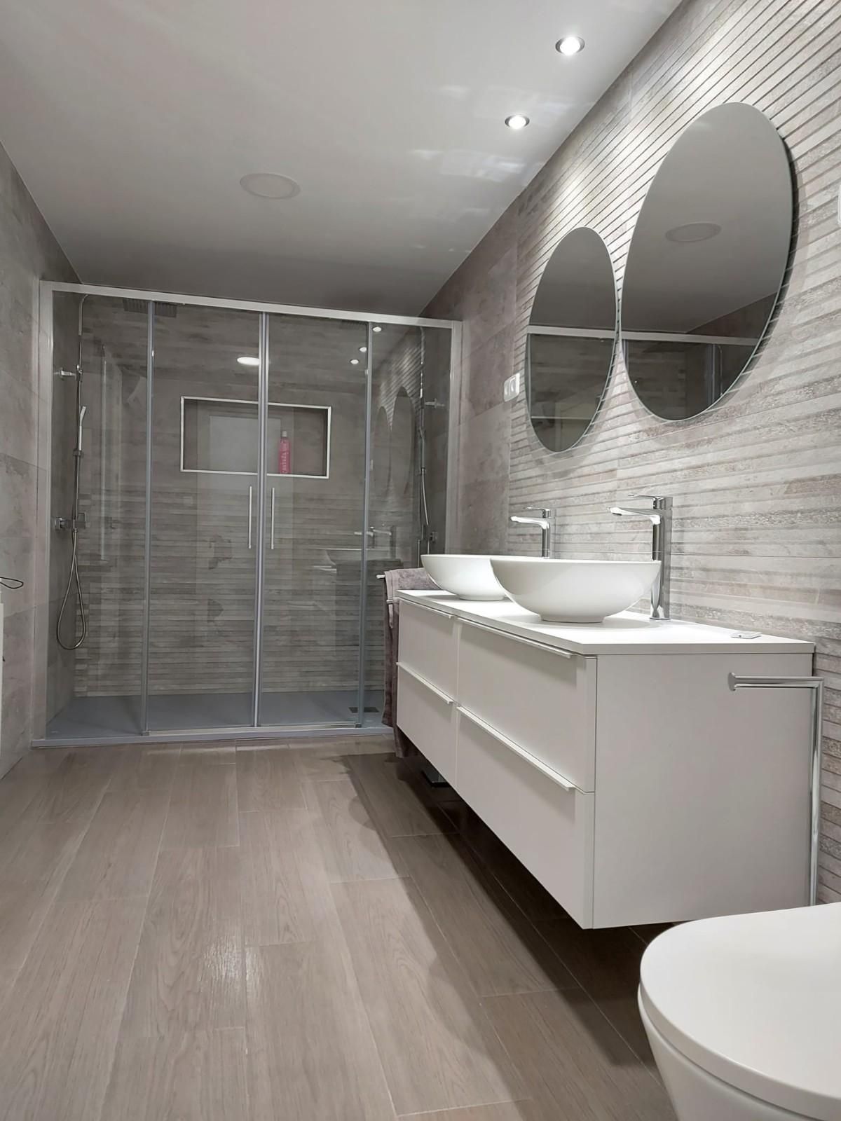Baño moderno con lavabo doble, espejos redondos, paredes alicatadas y mampara de ducha de cristal.