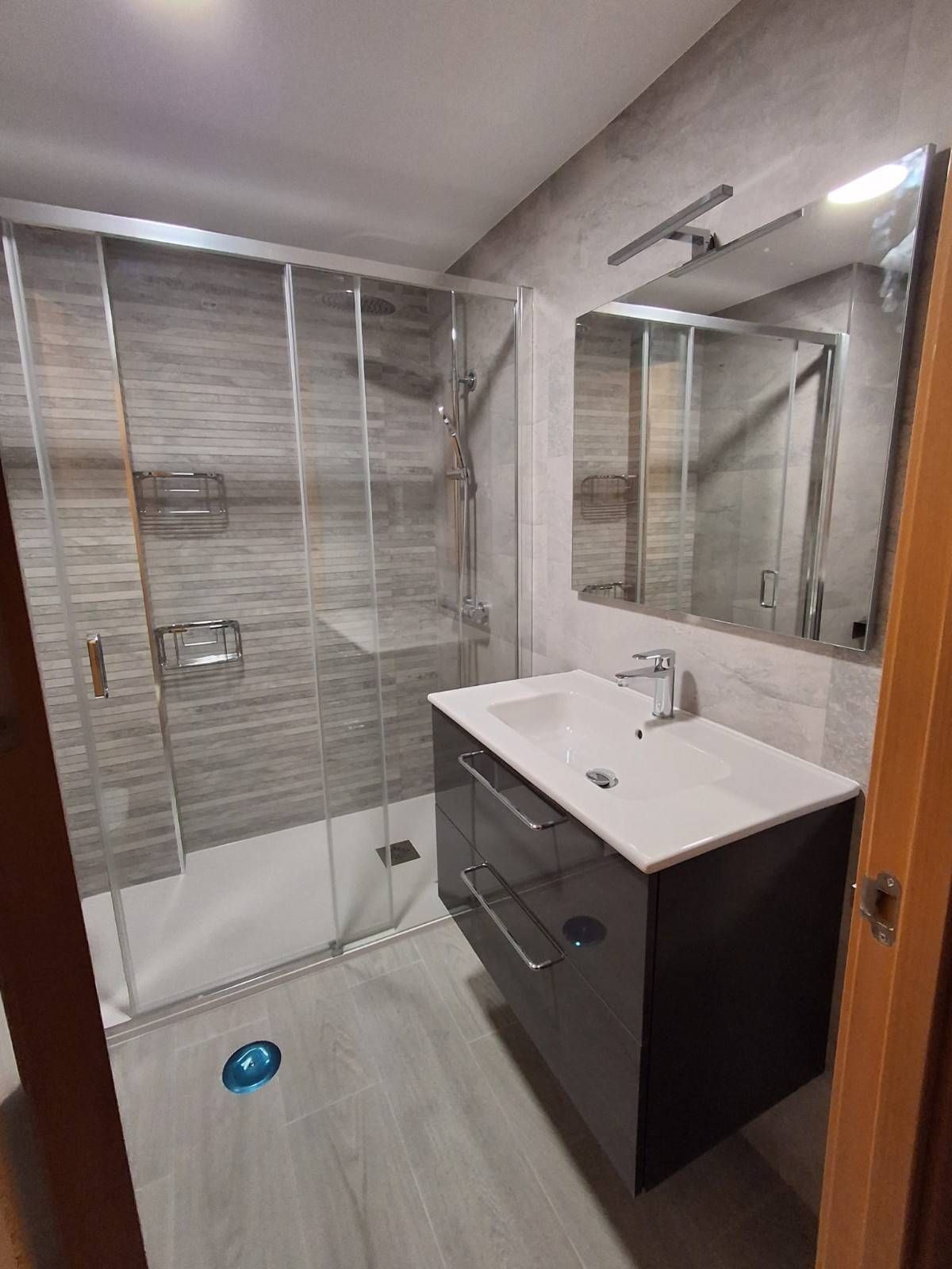 Baño moderno con ducha de azulejos grises, tocador oscuro, lavabo blanco y espejo grande.