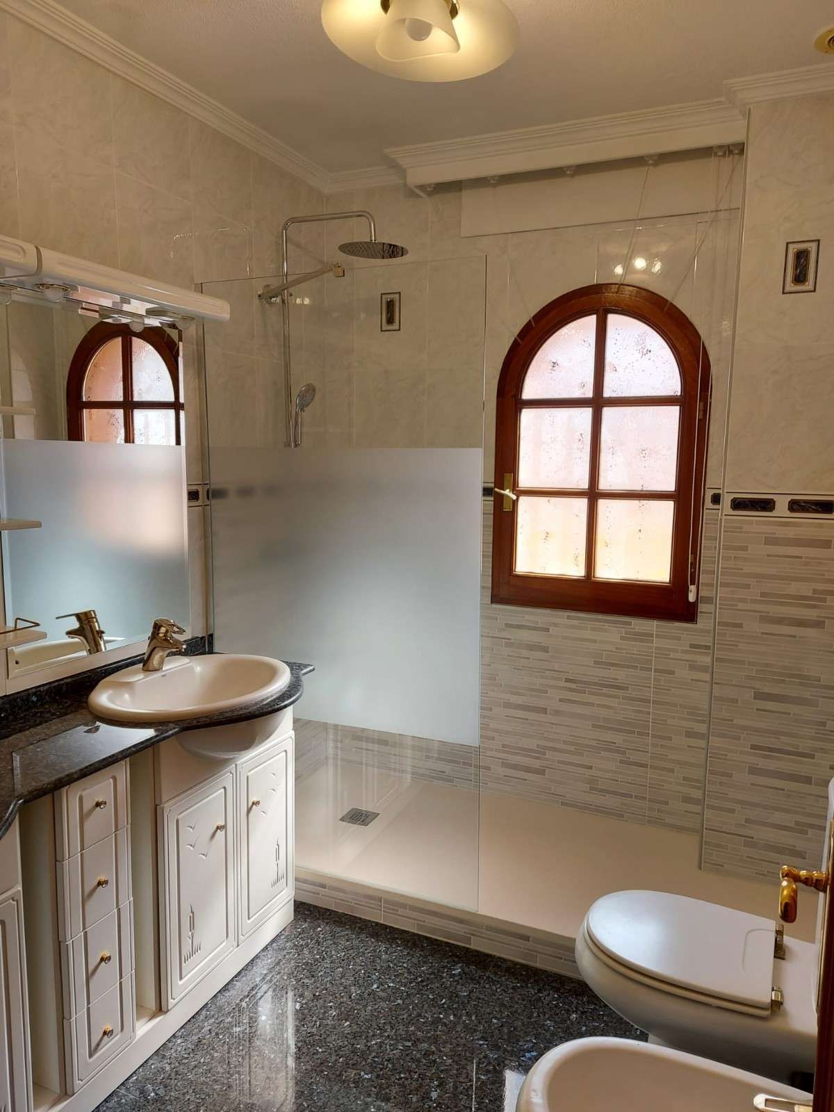Baño con paredes de azulejos blancos, ventanas de madera arqueadas, lavabo con tocador, ducha e inodoro.