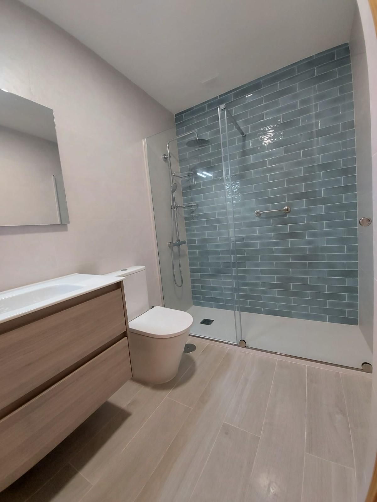 Baño moderno con tocador de madera, inodoro y ducha con mampara de cristal y pared de azulejos azules.