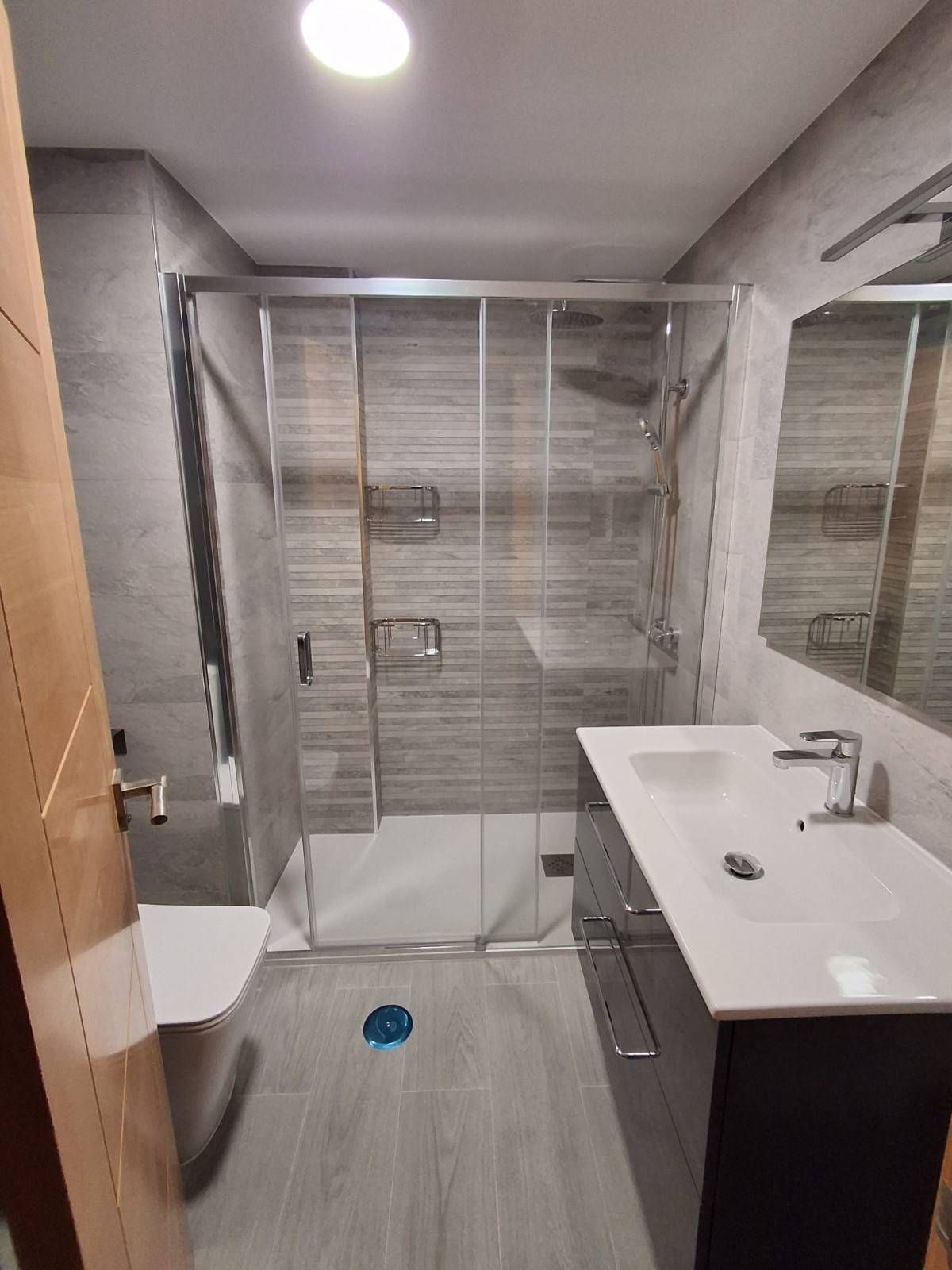 Baño moderno con ducha de cristal, espejo de pared, lavabo blanco y suelo de baldosas grises.