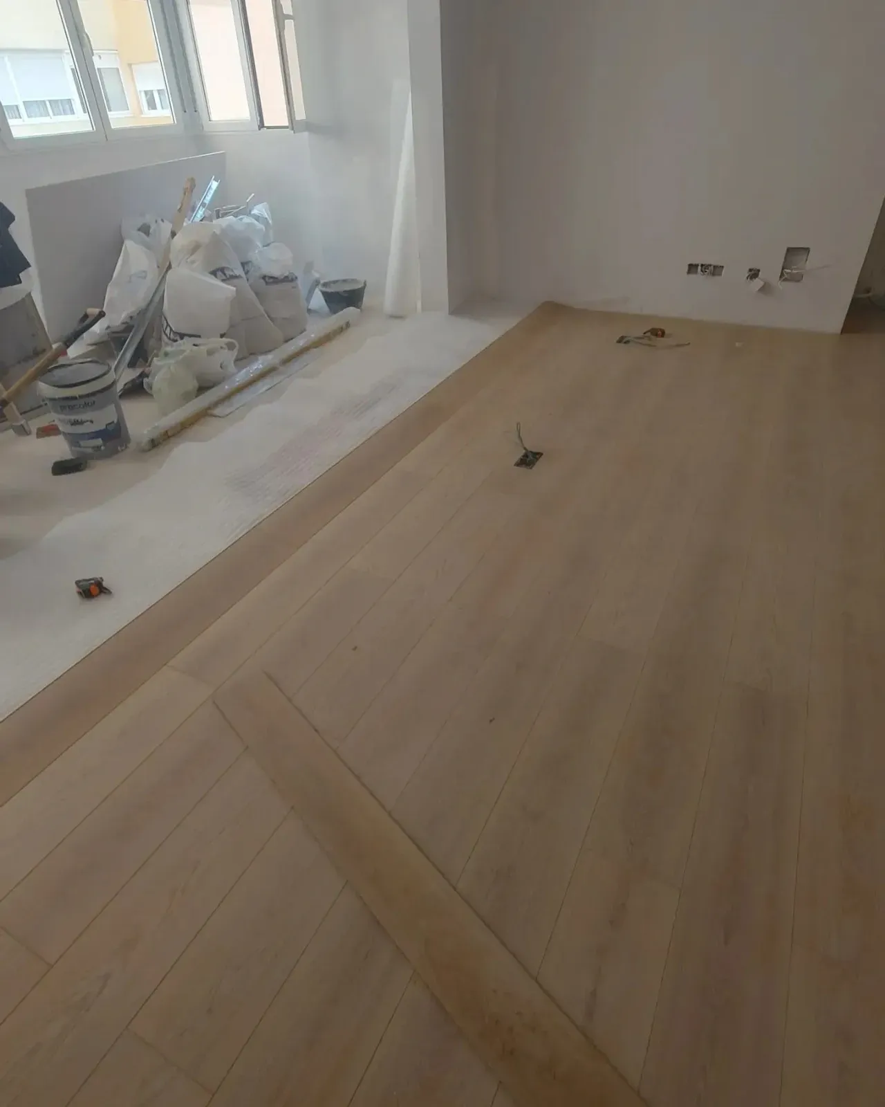 Habitación parcialmente renovada con suelo de madera clara, herramientas y escombros cerca de las ventanas.