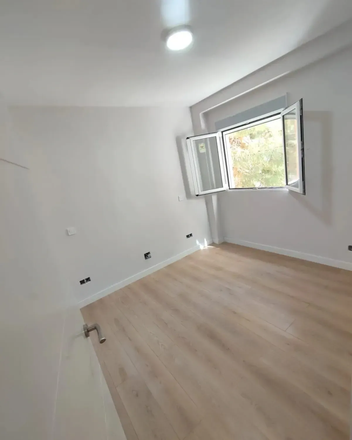 Habitación blanca vacía con suelo de madera clara y ventana abierta que deja entrar la luz del día.