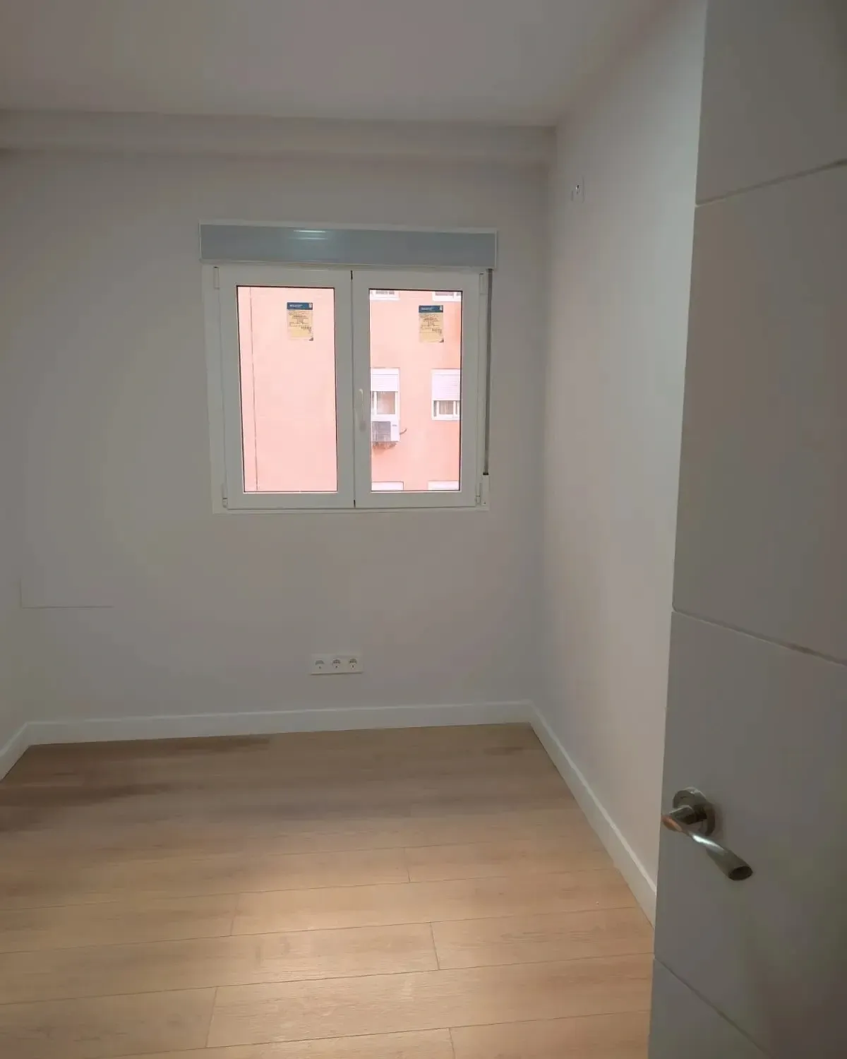 Habitación blanca vacía con suelo de madera, una ventana y una puerta entreabierta.