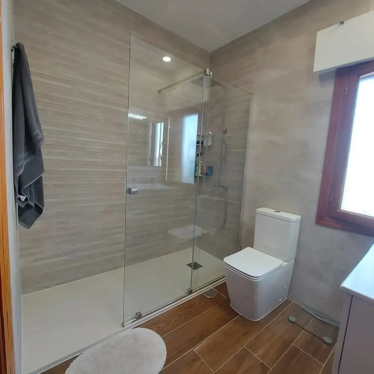 Baño moderno con ducha de cristal, inodoro suspendido, suelo de madera y luz de ventana.