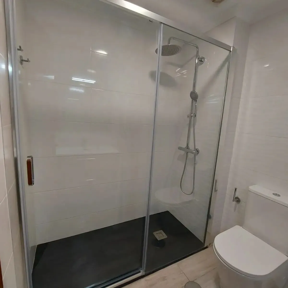 Baño moderno con ducha a ras de suelo de cristal, azulejos blancos y un inodoro.
