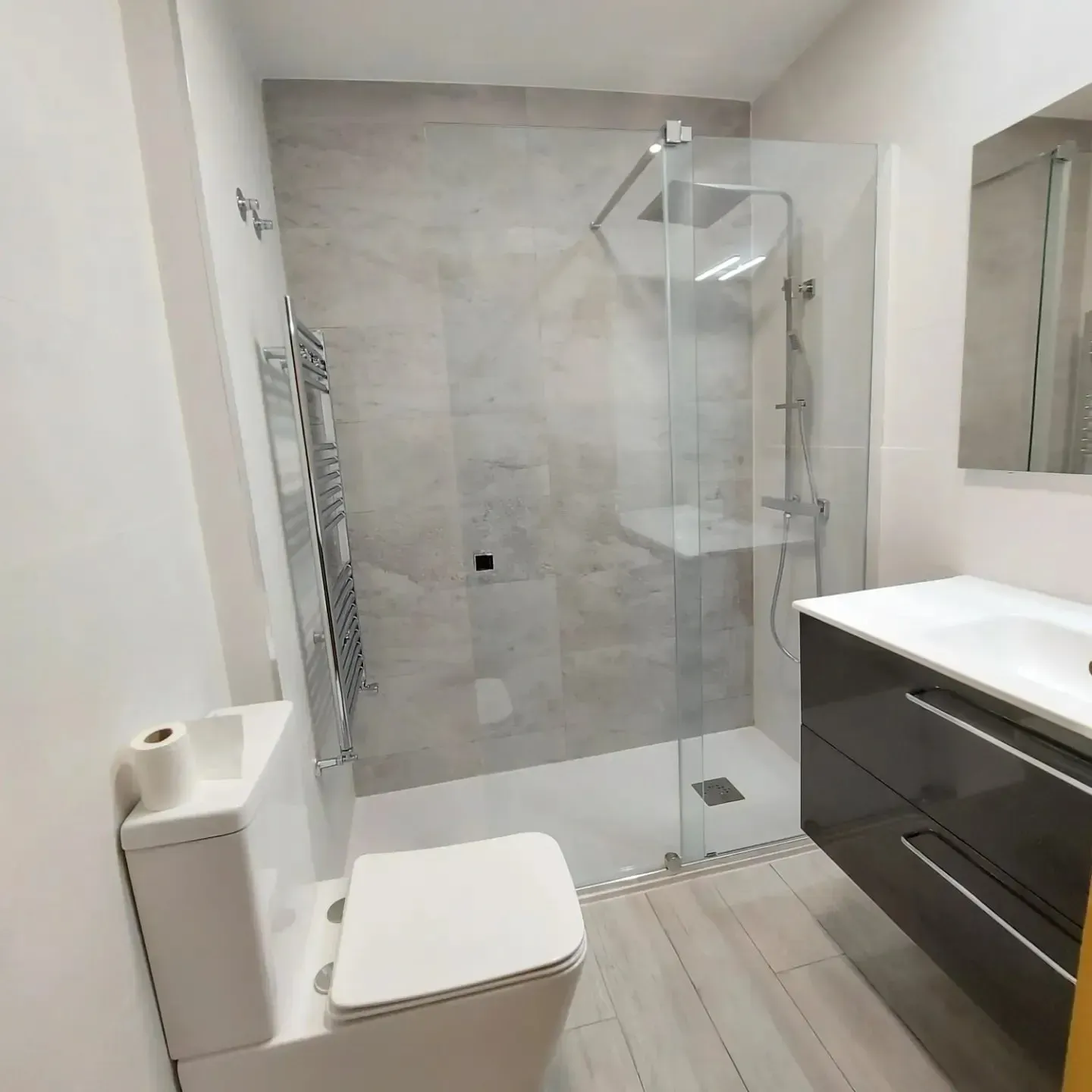 Baño moderno con inodoro, ducha de cristal y lavabo con mueble oscuro.