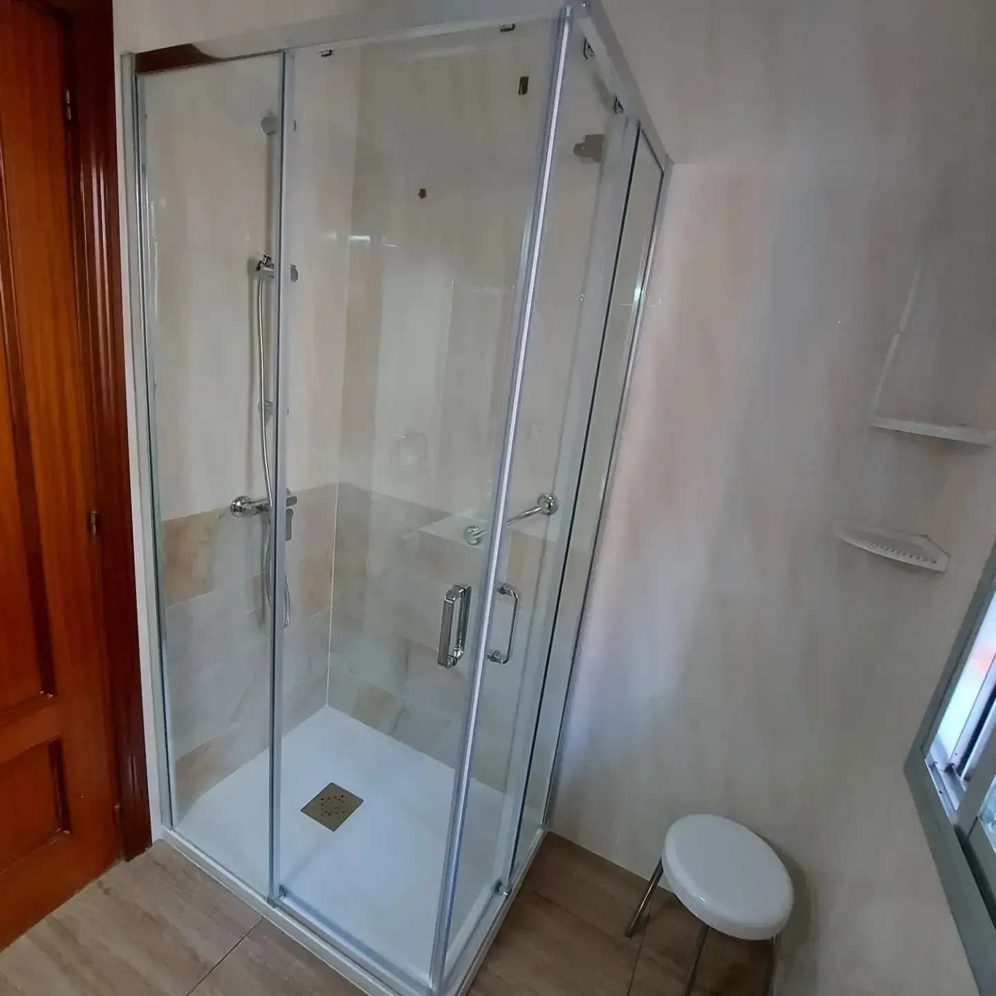 Cabina de ducha de esquina con puertas de cristal transparente, azulejos blancos y un pequeño taburete en un baño.