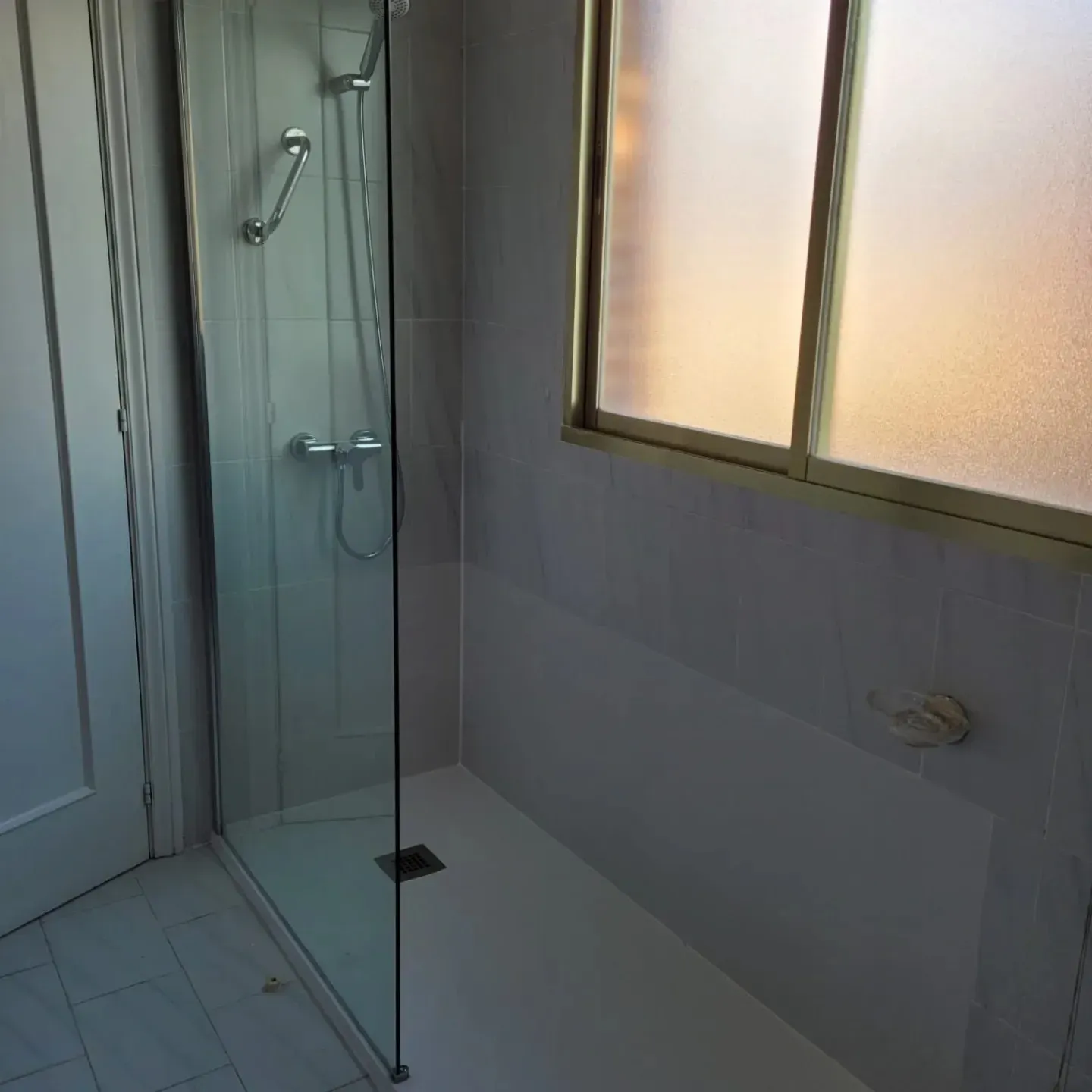 Baño pequeño con cabina de ducha de cristal, suelo de baldosas y ventana esmerilada a la derecha.