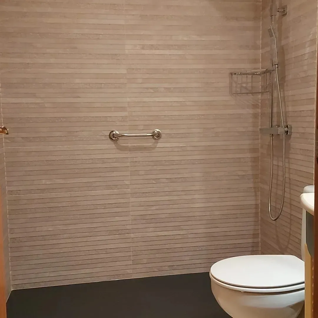 Baño pequeño con azulejos beige, barra de apoyo en la pared, mampara de ducha de cristal e inodoro blanco.