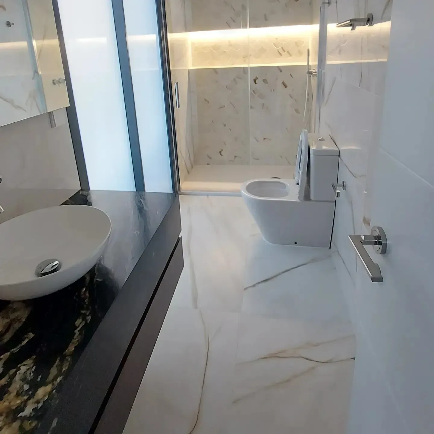 Baño moderno con azulejos de mármol blanco, ducha de cristal, inodoro suspendido y lavabo con mueble negro.