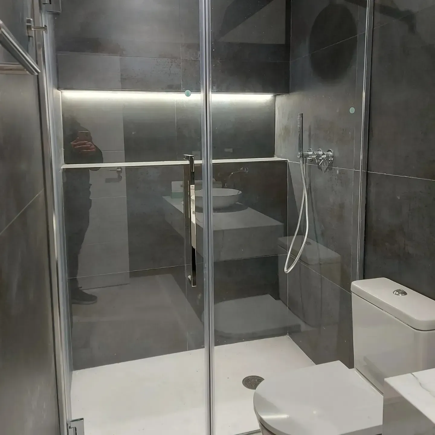 Baño moderno con cabina de ducha de cristal, inodoro y lavabo sobre paredes alicatadas en tonos oscuros.
