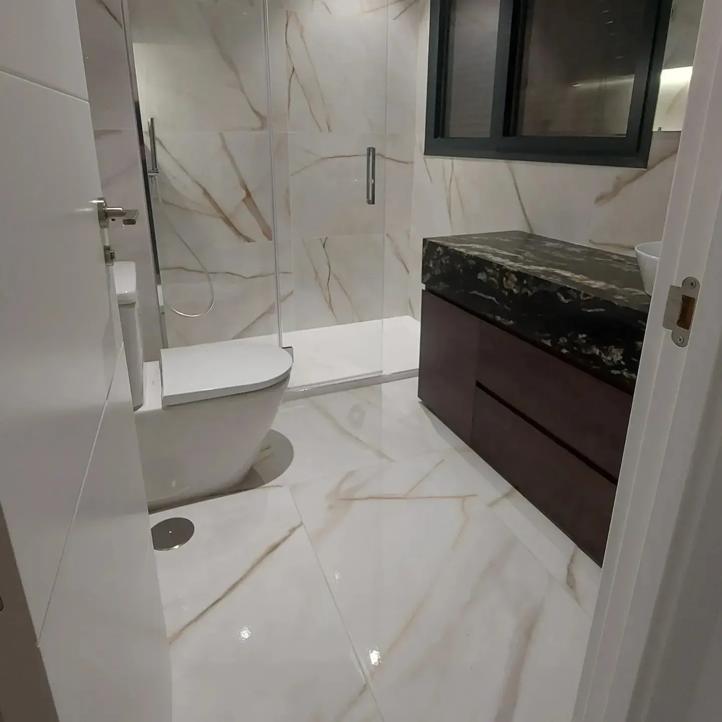 Baño moderno con azulejos de mármol blanco, tocador negro, bañera y ventana sobre el lavabo.