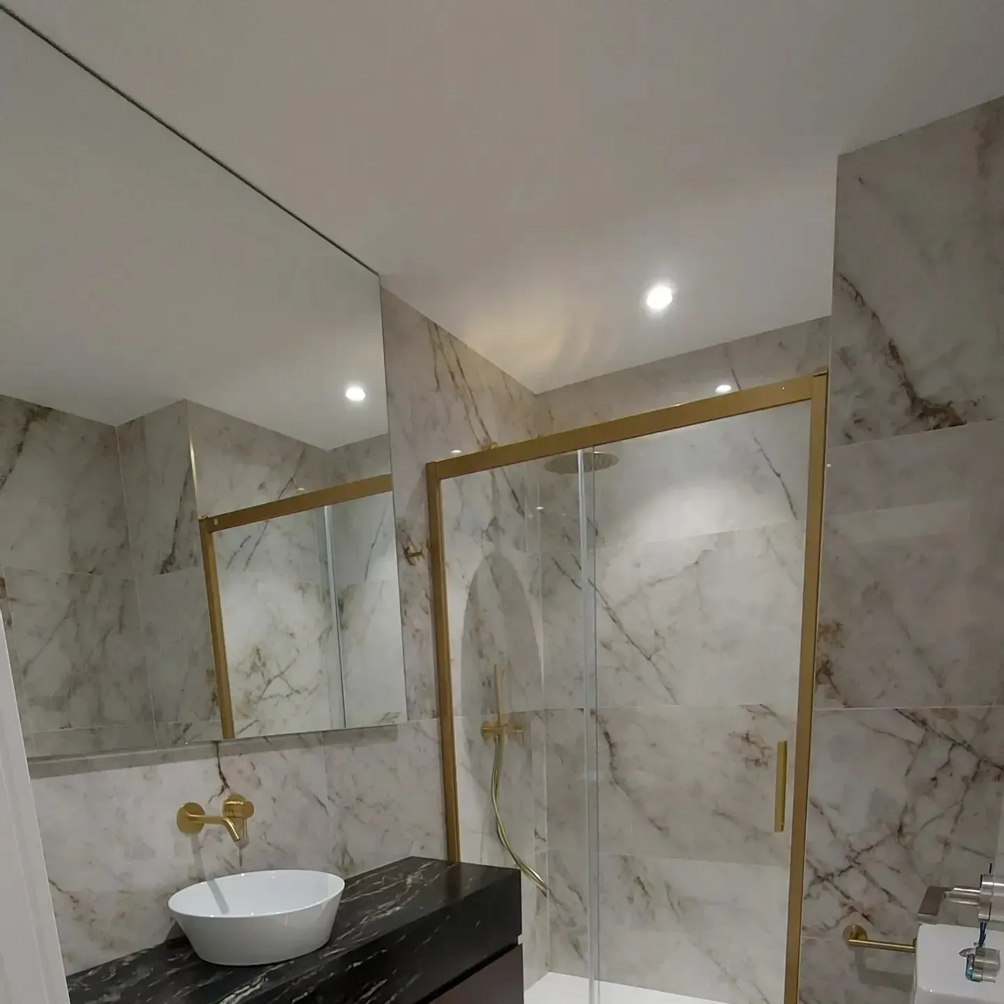 Baño moderno de mármol con ducha con marco dorado, lavabo blanco sobre encimera y pared con espejo.