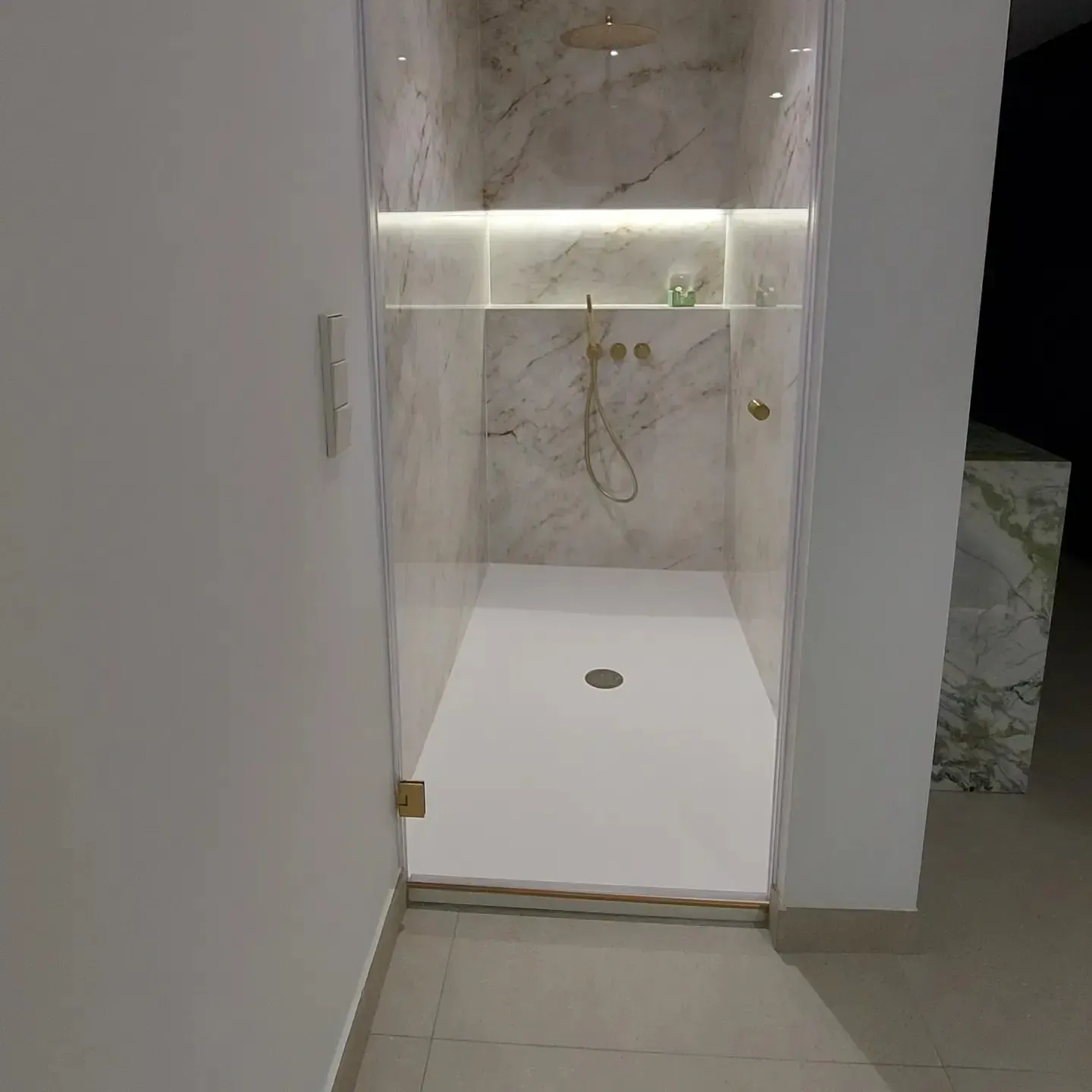 Ducha moderna a ras de suelo con azulejos de mármol, estante incorporado y grifería de latón, vista a través de una puerta.