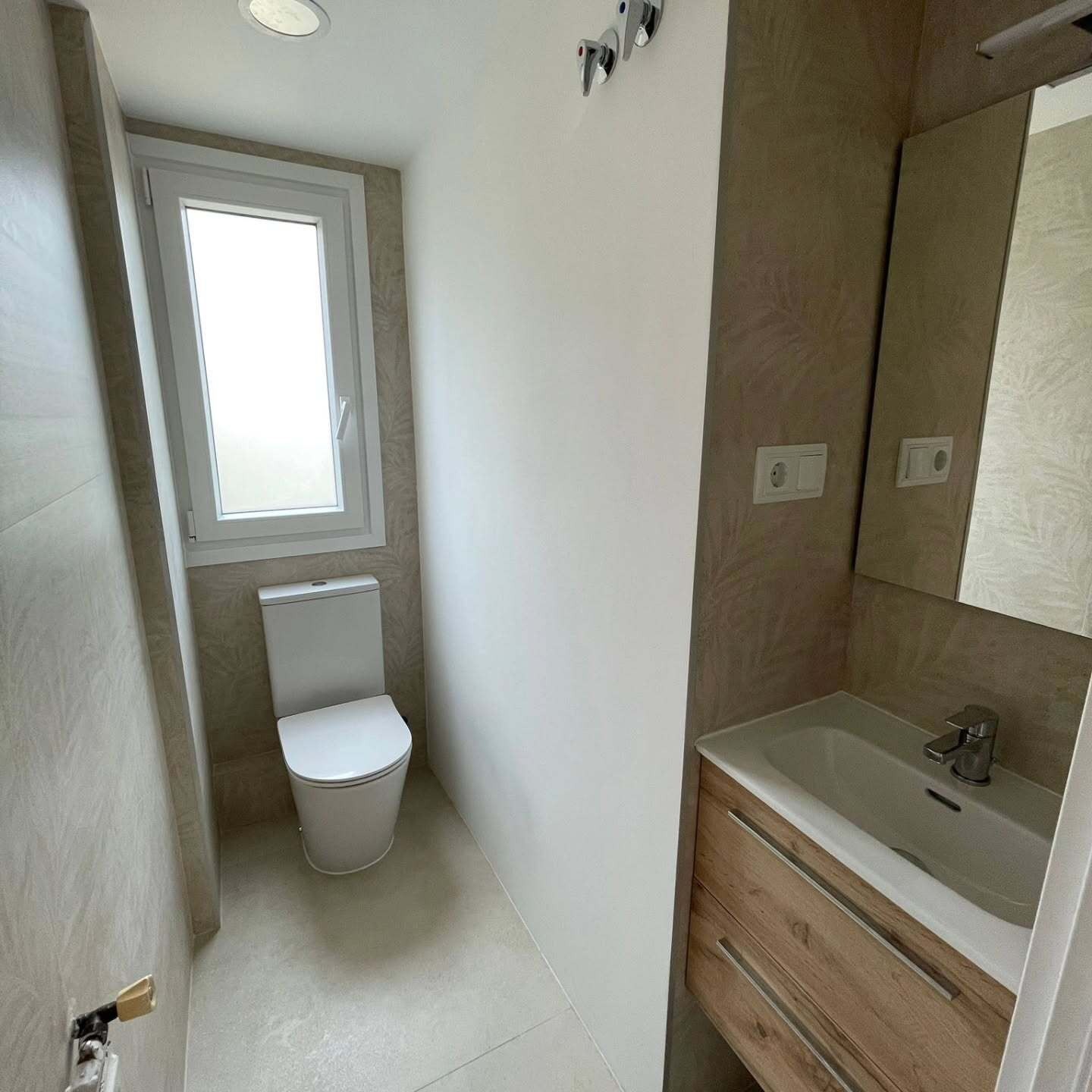 Baño moderno y estrecho con inodoro, ventana pequeña y tocador de madera con lavabo.