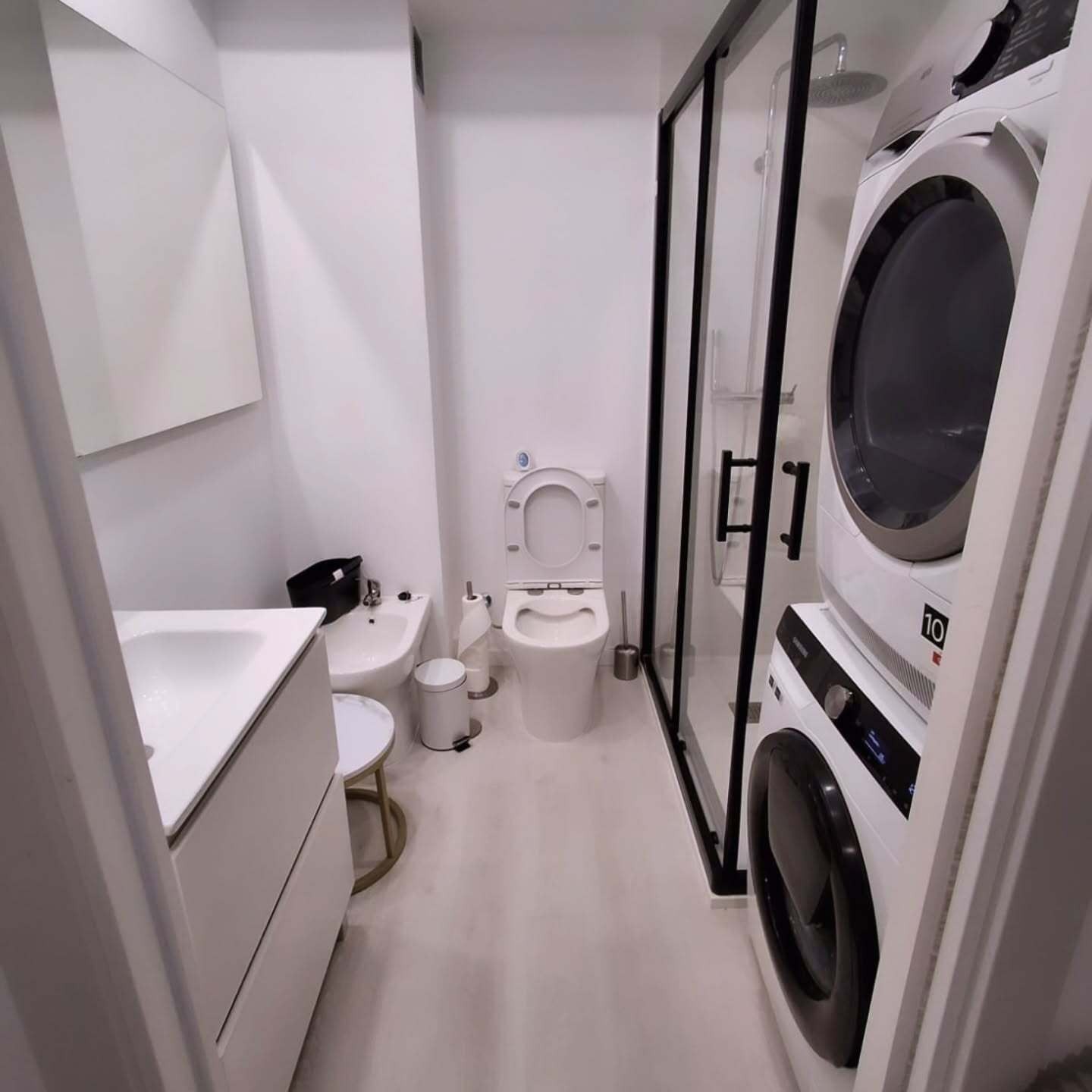 Baño estrecho de color blanco con lavadora y secadora apiladas, cabina de ducha, lavabo e inodoro.