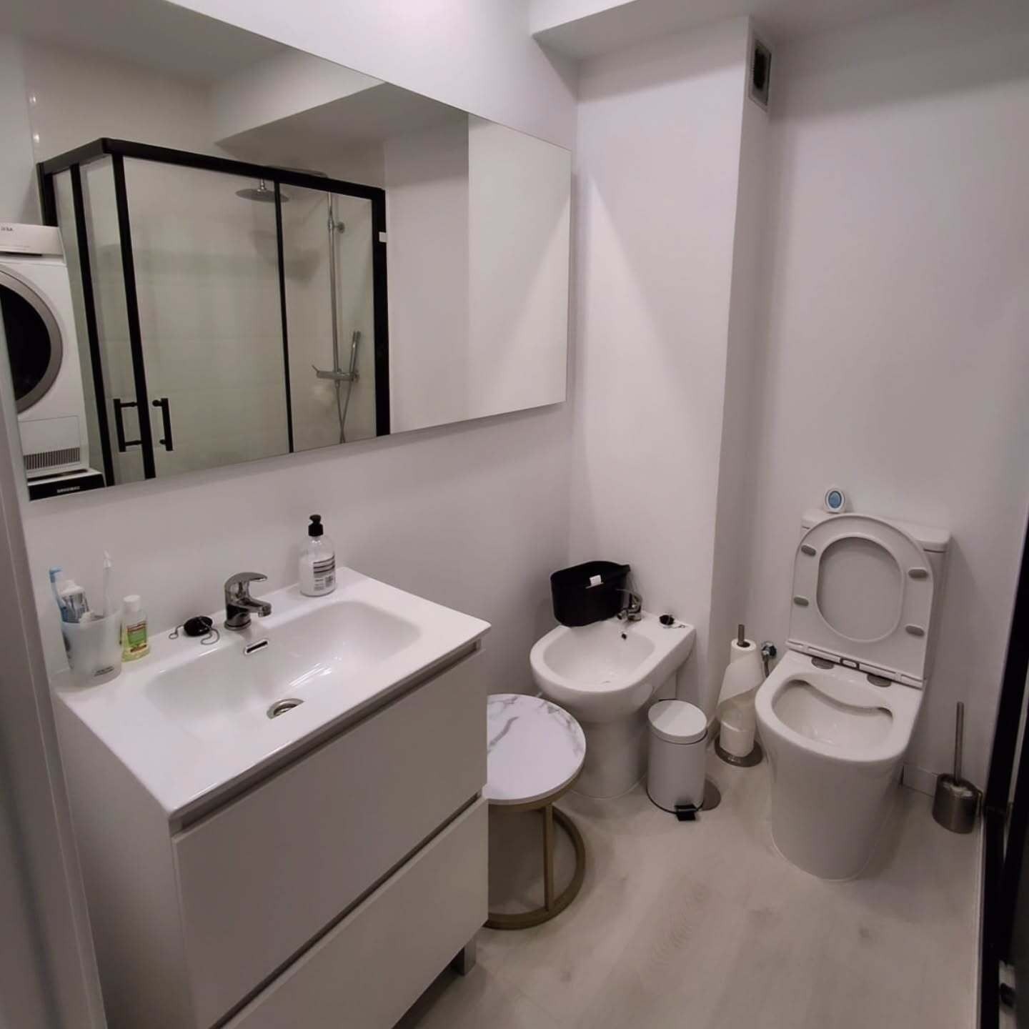 Baño moderno blanco con lavabo, espejo, bidé, inodoro y cabina de ducha de cristal.