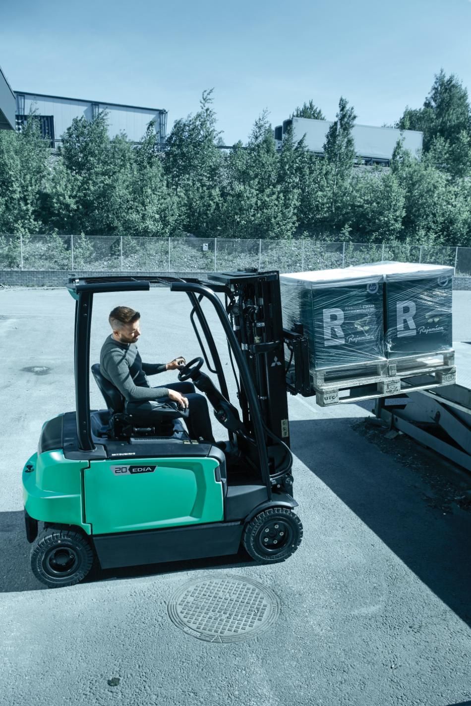Un cariste en chemise grise conduit un chariot élévateur vert à l'extérieur, transportant une palette de marchandises en cartons.