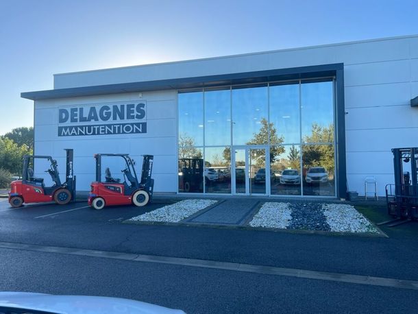 Un bâtiment commercial blanc portant l'enseigne DELAGNES MANUTENTION, avec deux chariots élévateurs rouges garés devant.