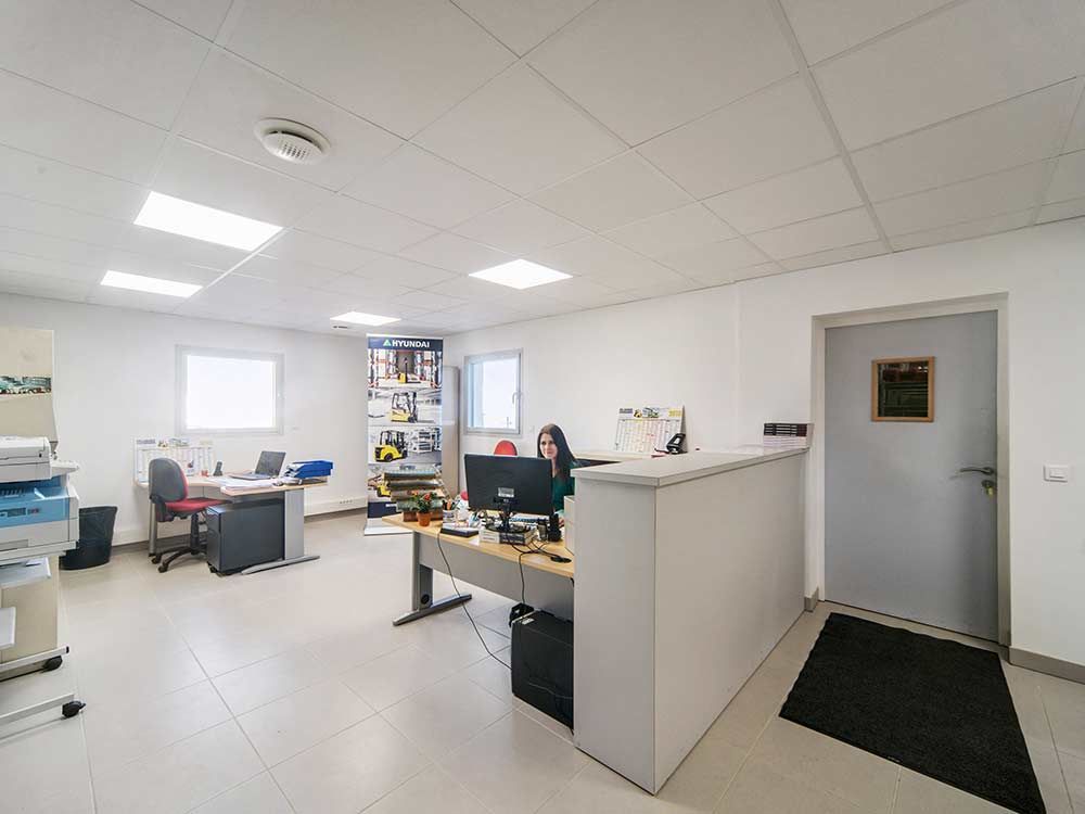 Intérieur d'un bureau avec un bureau, une personne travaillant sur un ordinateur, un présentoir et une porte grise sur la droite.
