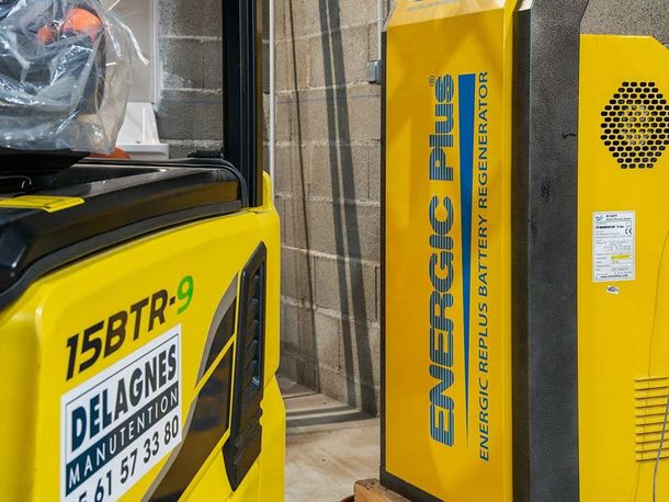 Un chariot élévateur jaune et un grand chargeur de batterie jaune Energic Plus sont côte à côte contre un mur de parpaings.