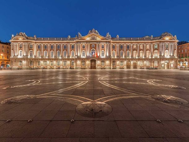 Le Capitole de Toulouse illuminé au crépuscule, devant une grande place pavée et ornée, à Toulouse, en France.