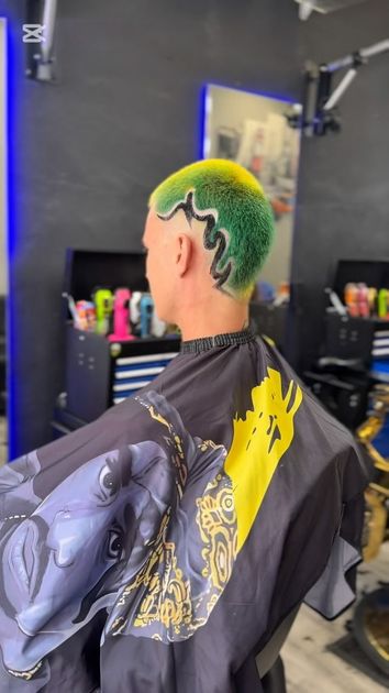 Persona con peinado verde y amarillo en una barbería, vistiendo una capa negra y amarilla.